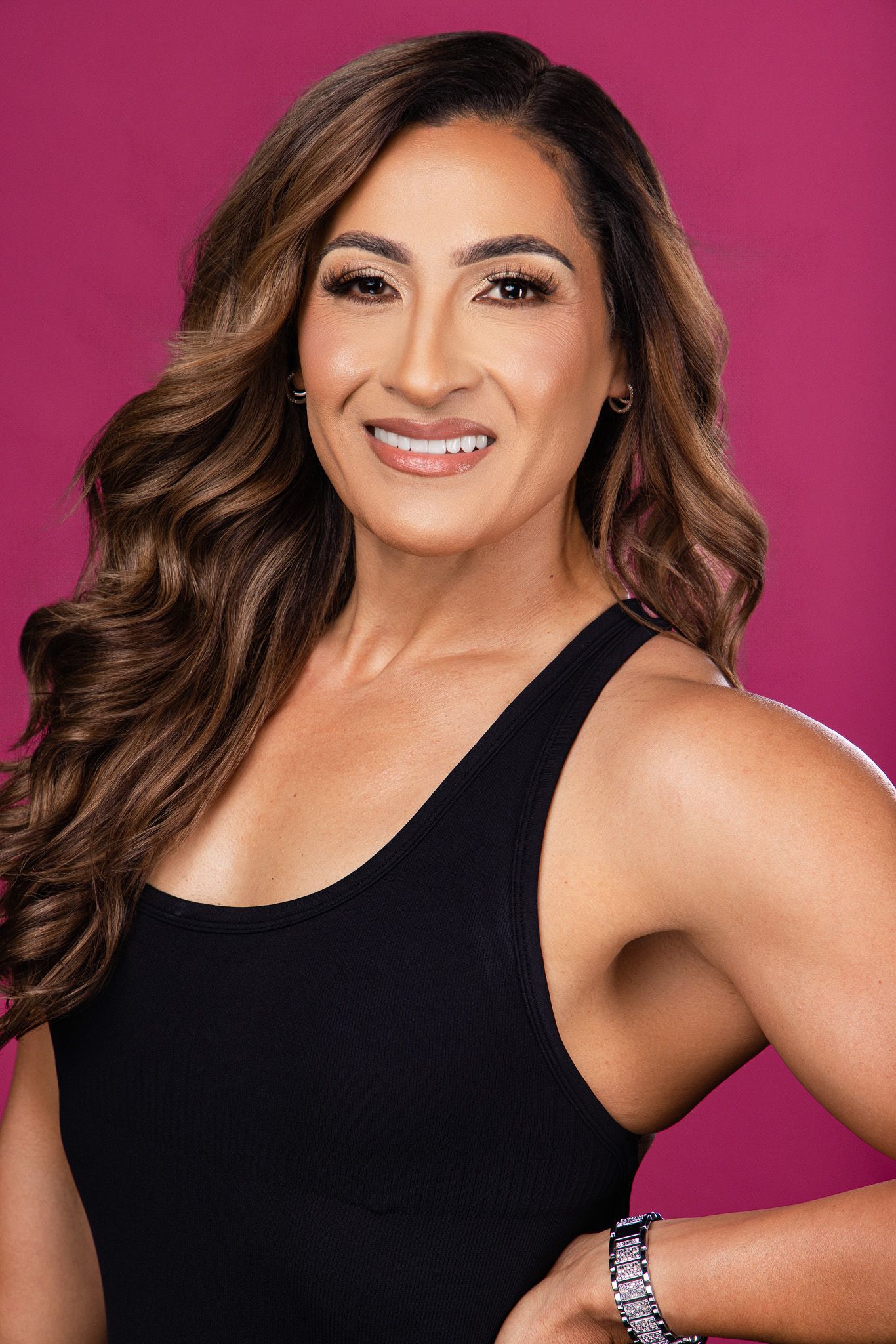 A confident woman with long wavy brown hair smiles in a black tank top against a vibrant pink background.