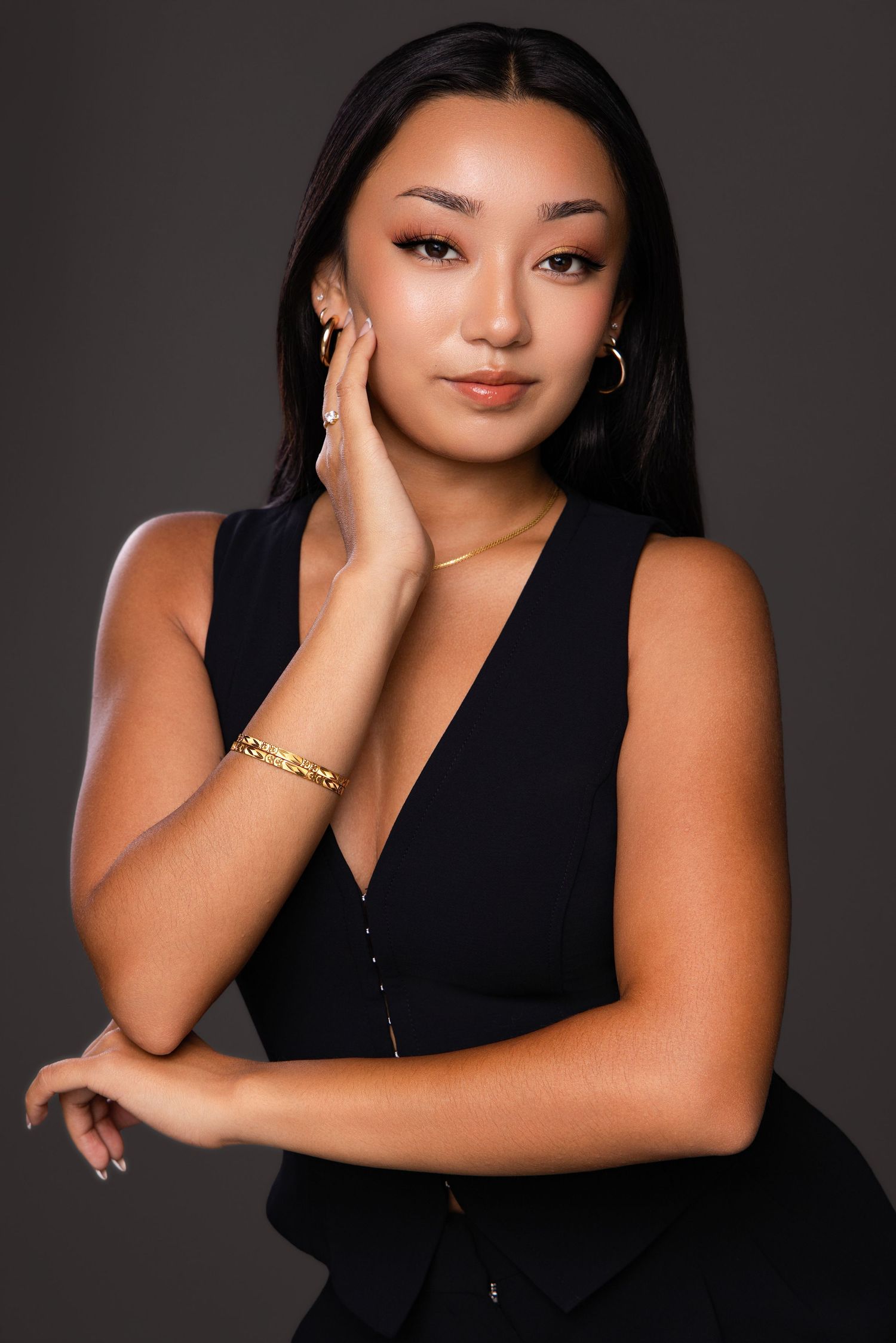Young woman in black sleeveless top with gold jewelry poses confidently against gray background.