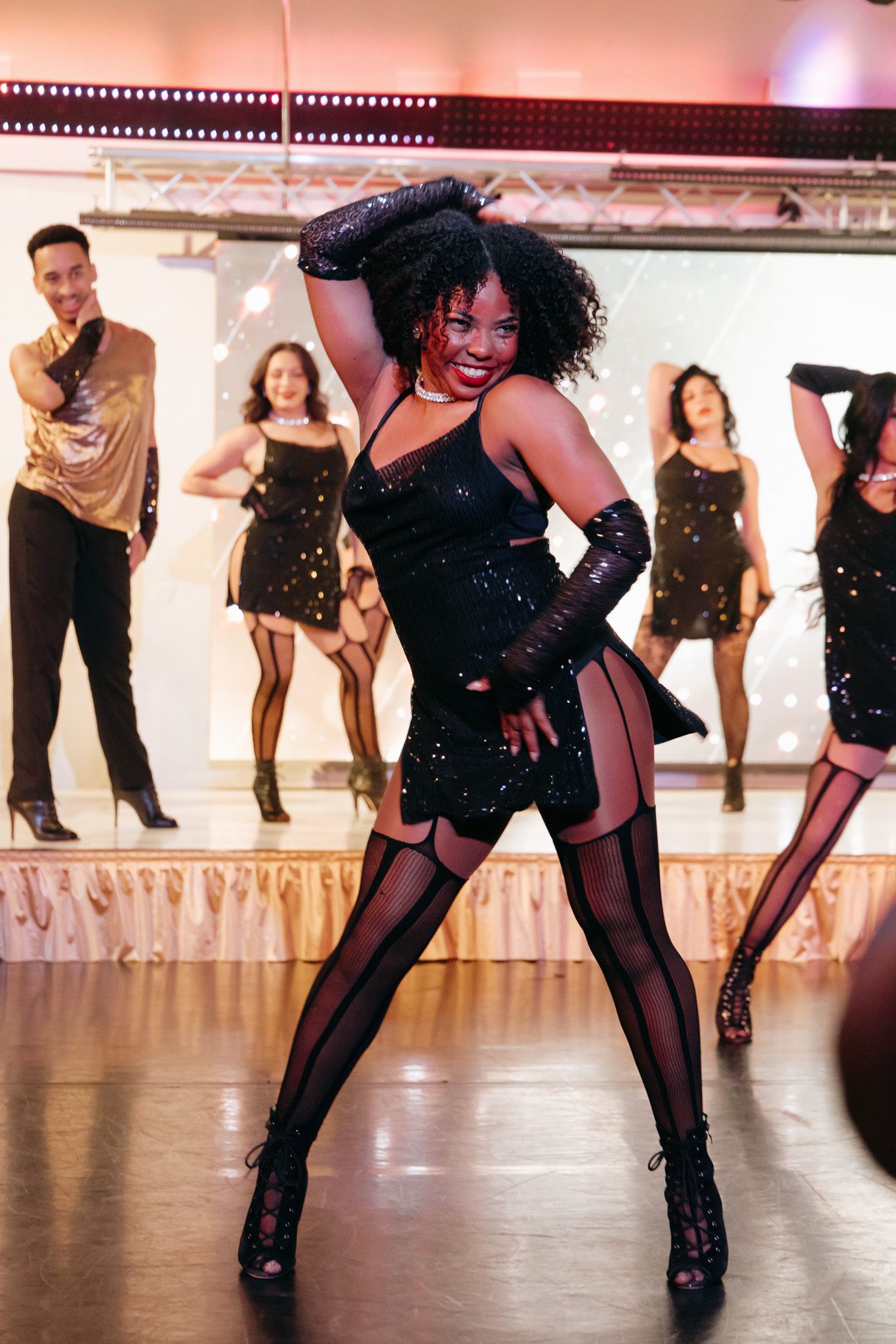Dancers perform a synchronized leg kick line in black costumes and stockings on a reflective dance studio floor.