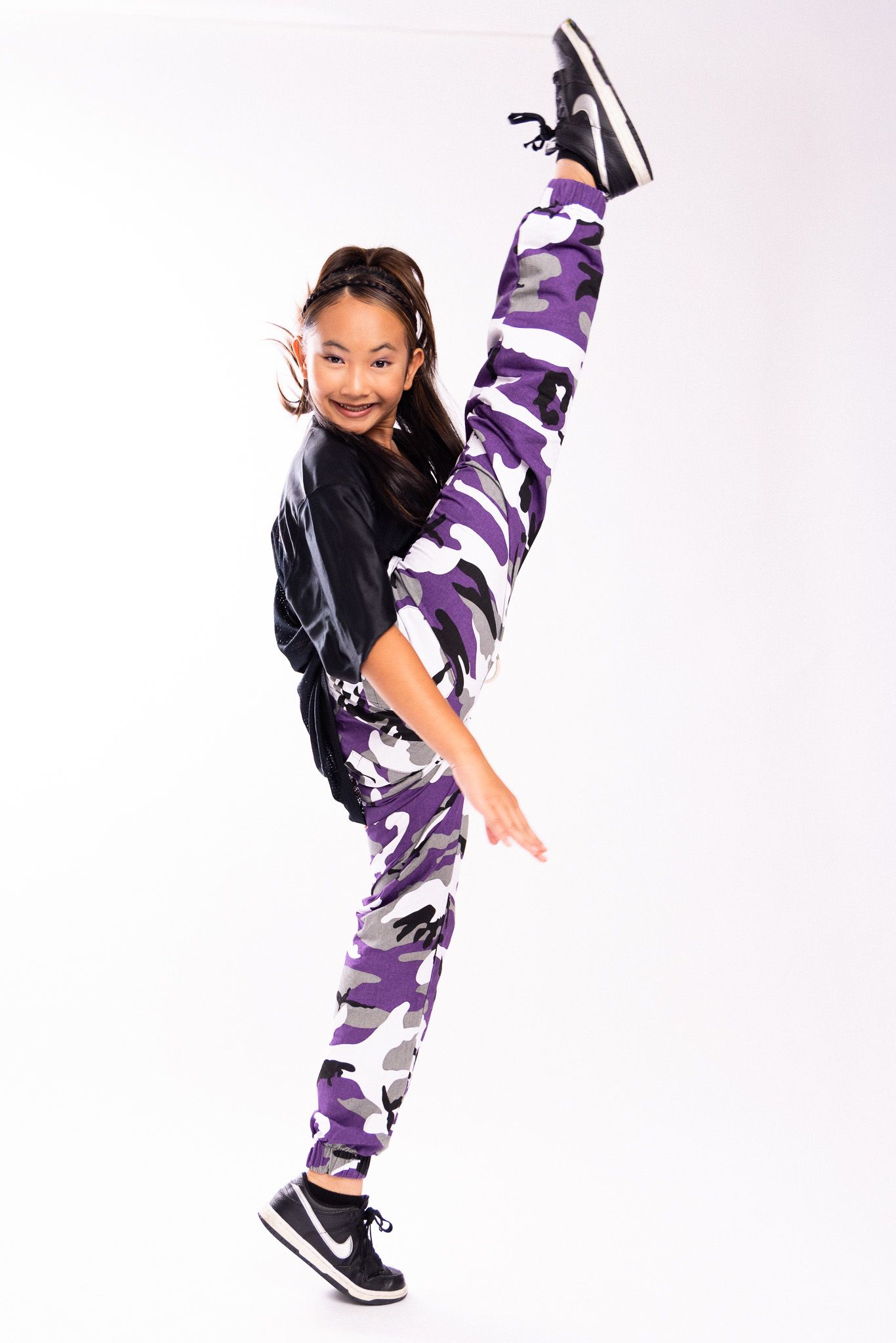 Person in purple camouflage pants and black top strikes a high kick dance pose against white background.