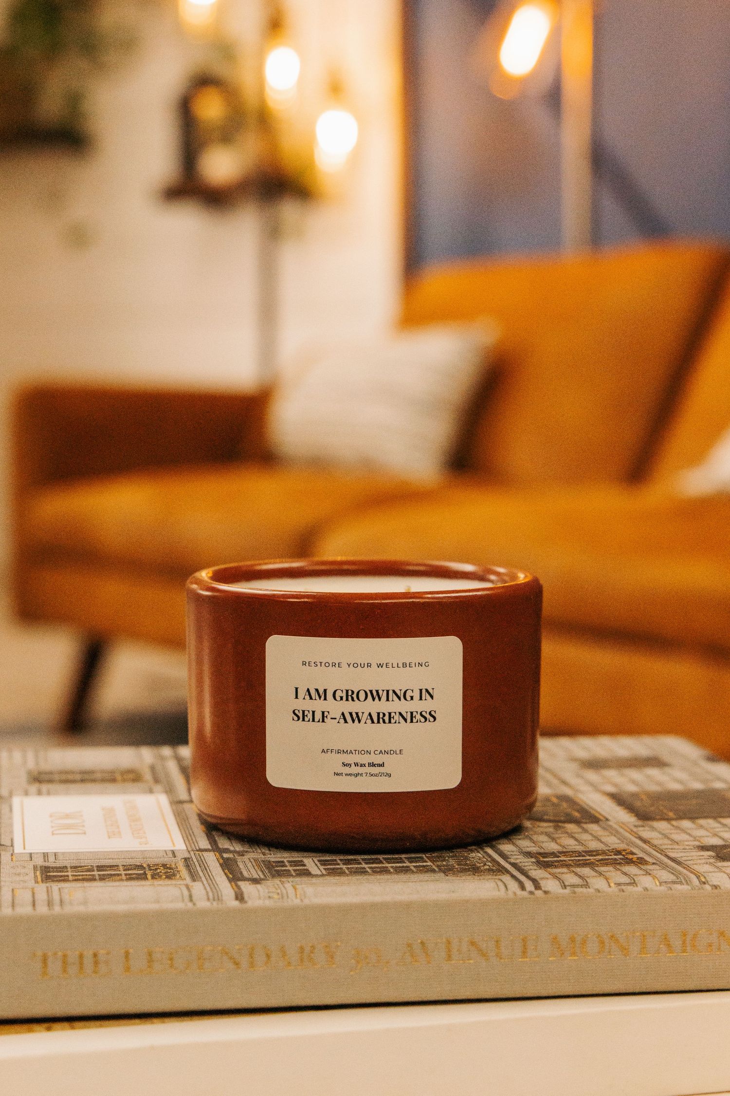 A brown scented candle with 'I am growing in self awareness' label displayed on a book with ambient lighting.