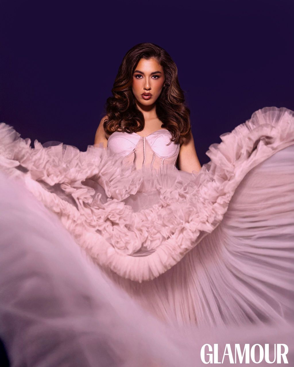 Fashion model in flowing pink ruffled gown poses dramatically against dark background for Glamour magazine.