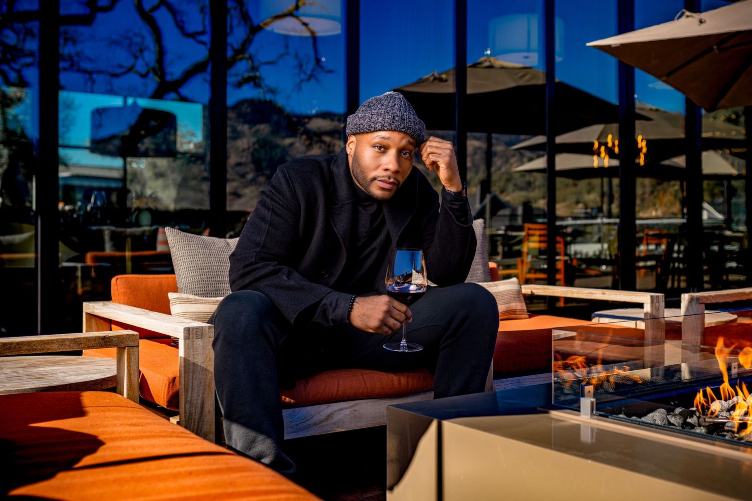 Man in beanie and black coat sits outdoors on orange cushioned furniture holding a drink near a fire pit.