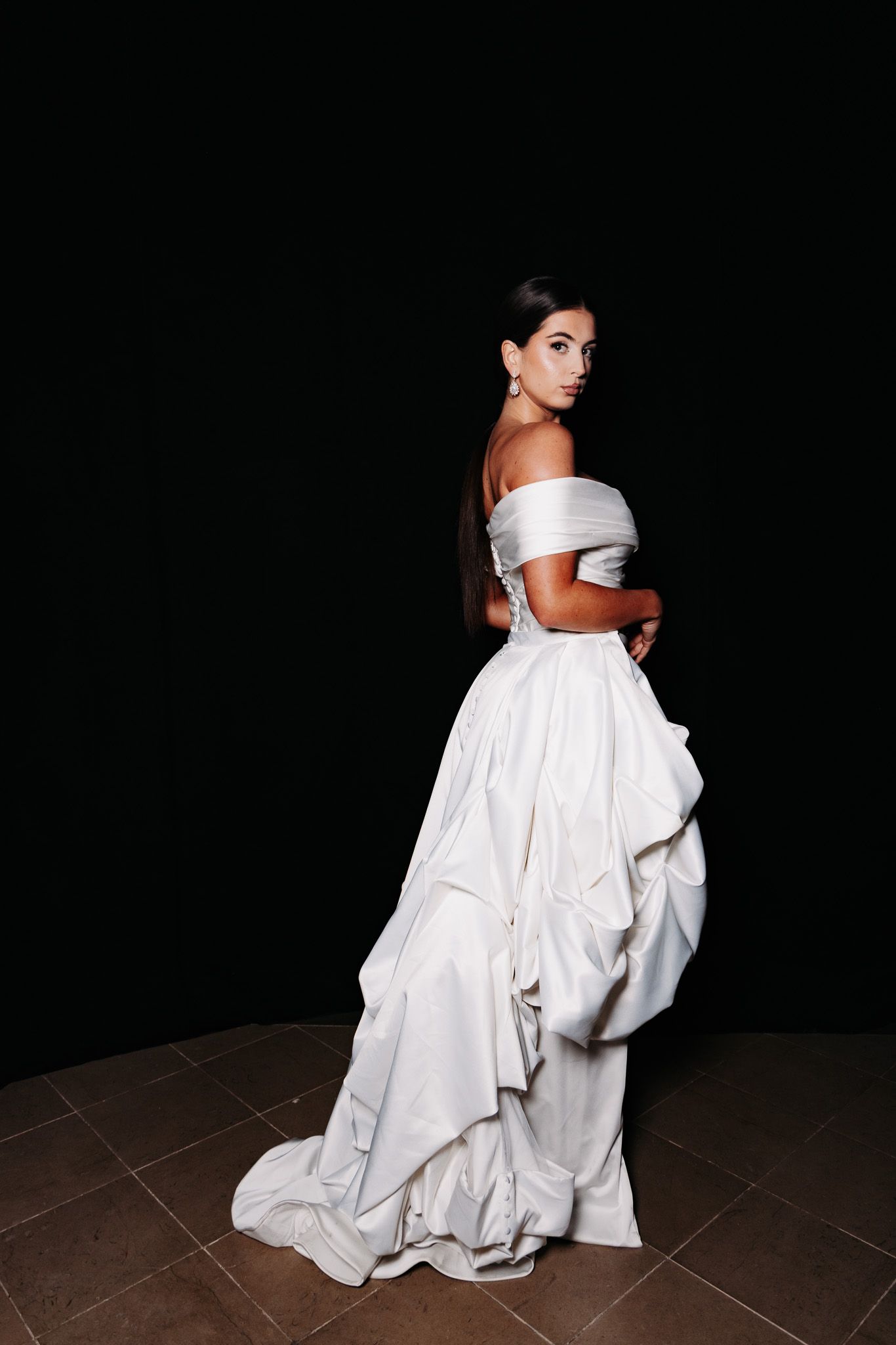 Elegant off-shoulder white ballgown with dramatic train captured in profile against dark background.