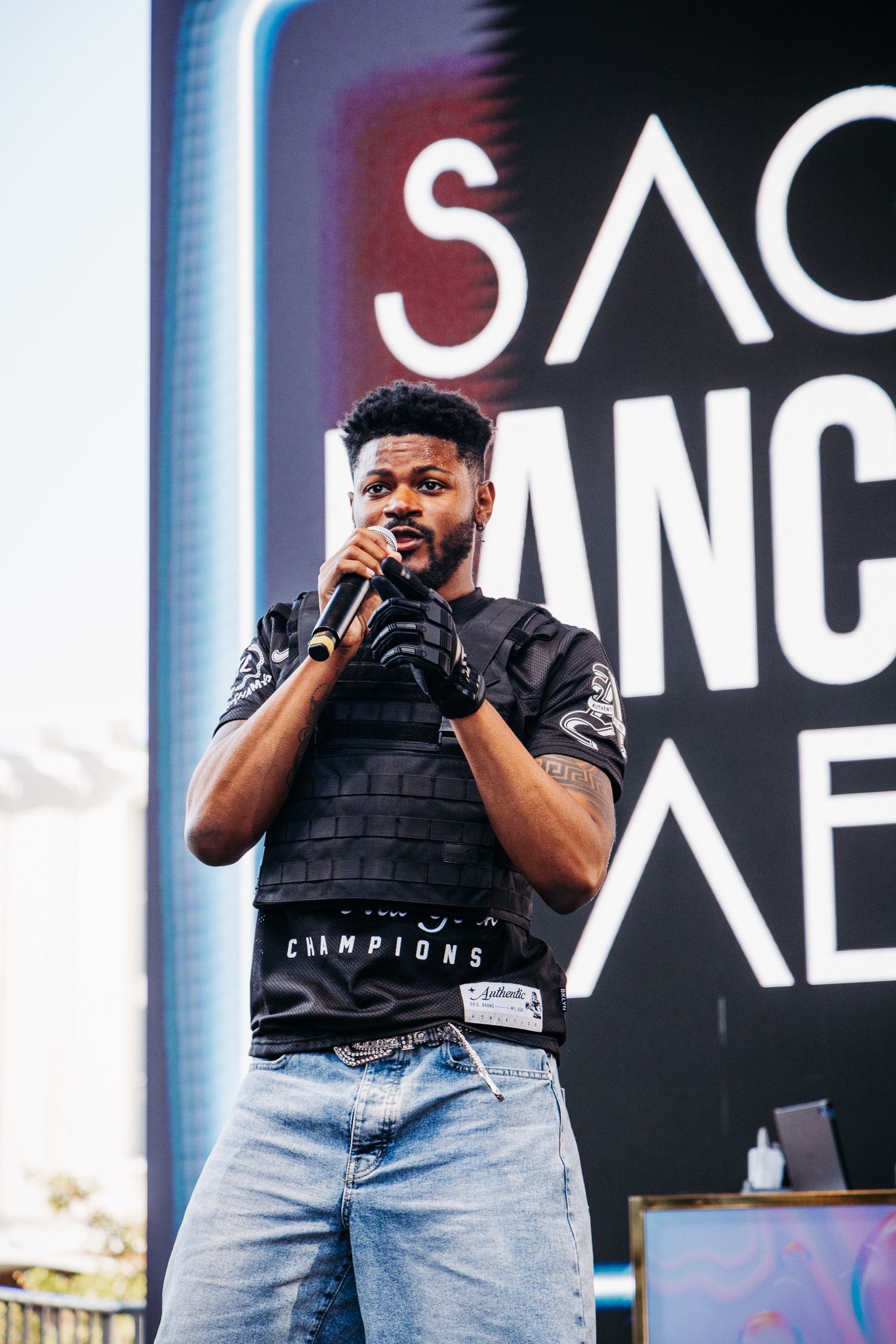 A performer in a black t-shirt and jeans sings passionately on stage with dramatic lighting and screens in the background.