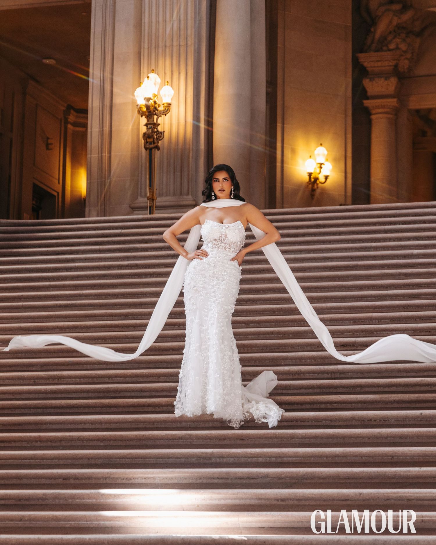 A bride in a white gown poses elegantly on grand marble steps inside an ornate building with glowing wall sconces.