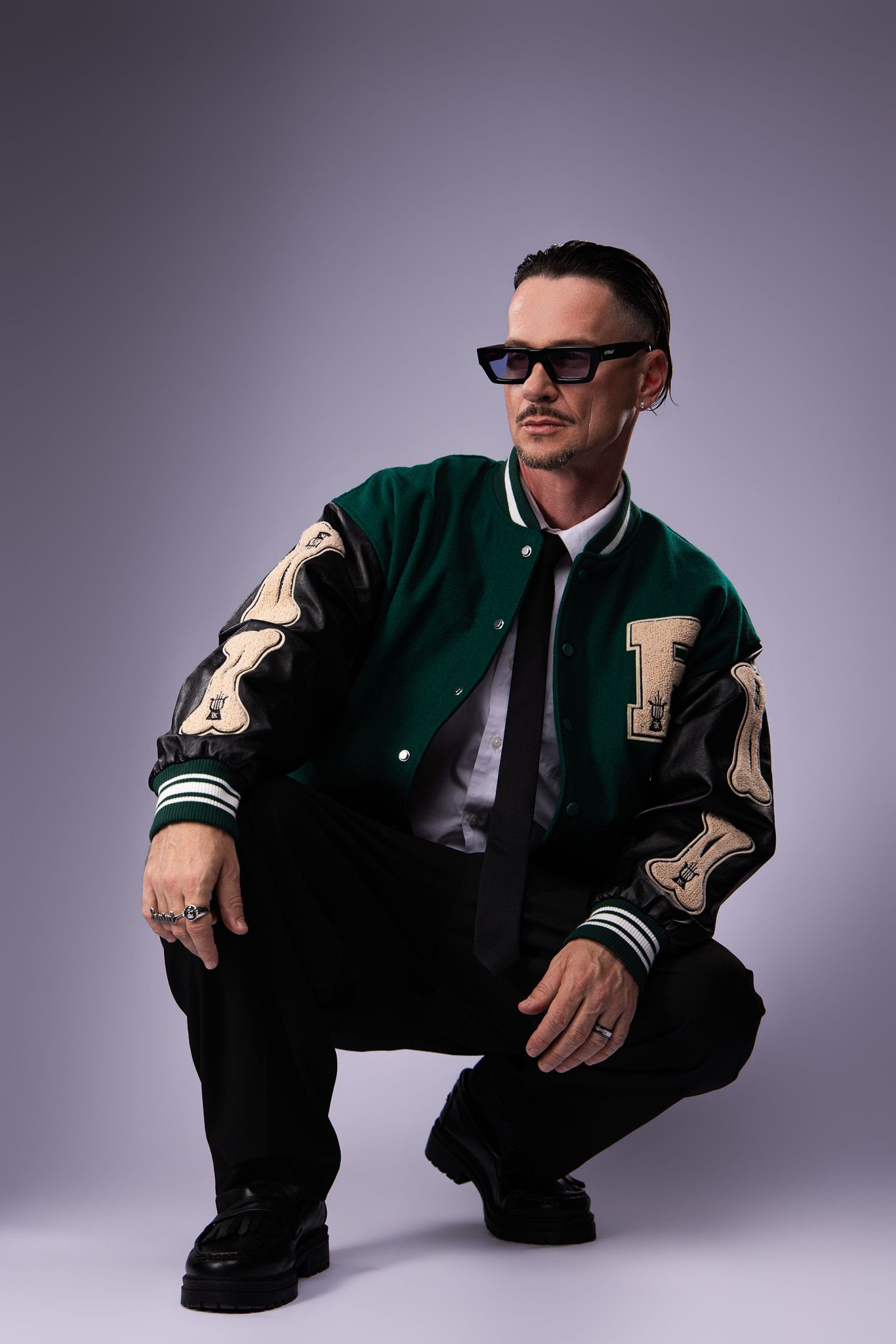Person in varsity jacket and sunglasses crouches in dynamic poses against gray studio backdrop.