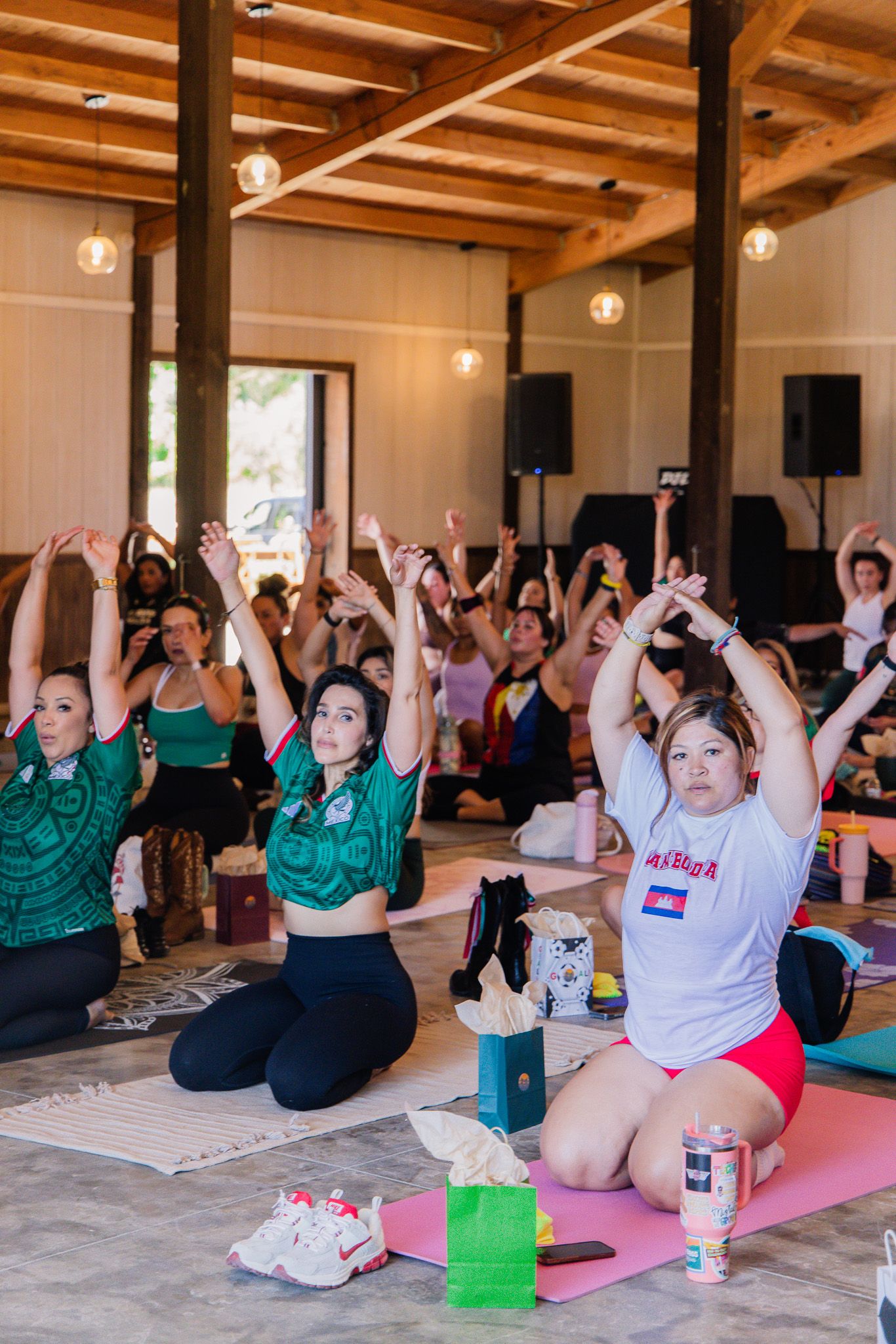 Group of women practicing yoga in a bright studio, sitting on mats with arms raised overhead.