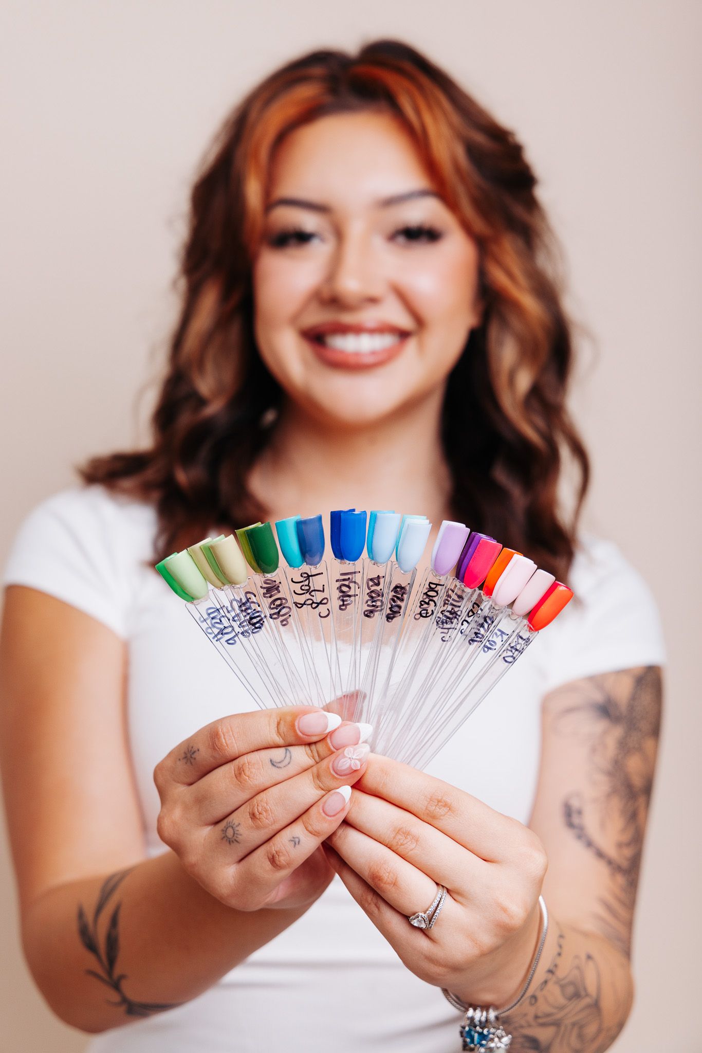 Smiling woman with tattoos holding a fan of colorful nail polish swatches in both hands.