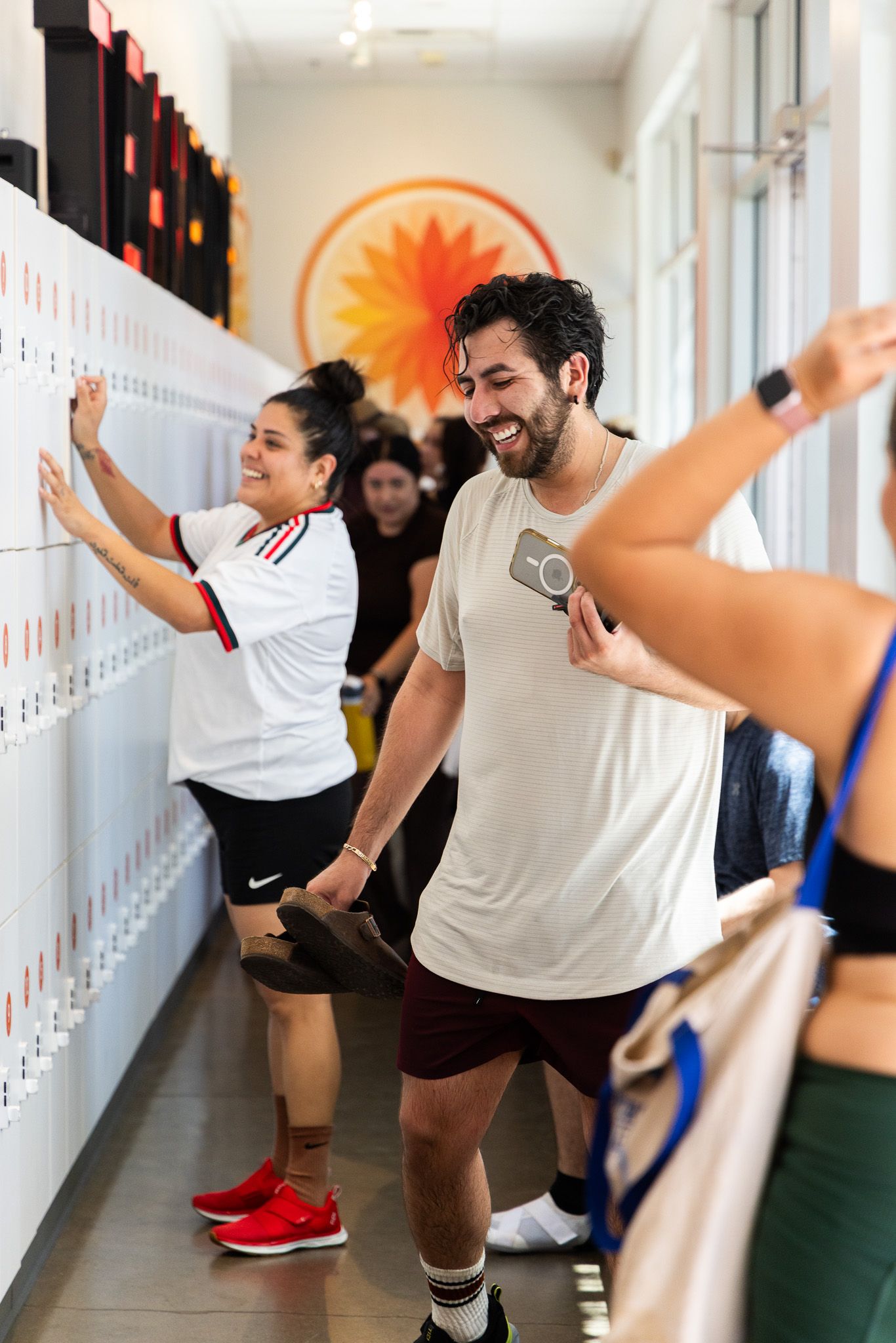 Two smiling gym-goers stretch and prepare near lockers in a bright fitness studio with a sunburst mural.