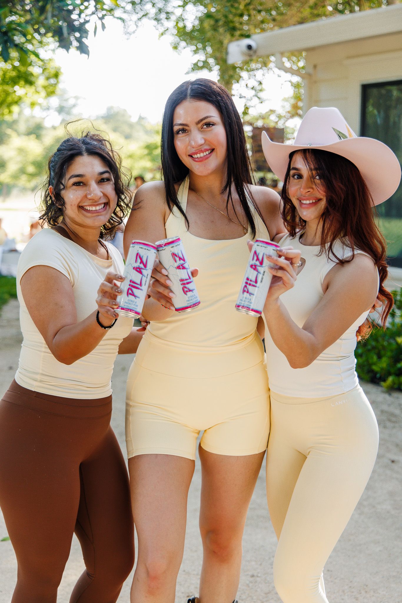 Three smiling women in coordinating outfits holding PILAZ canned drinks outdoors in sunny setting.