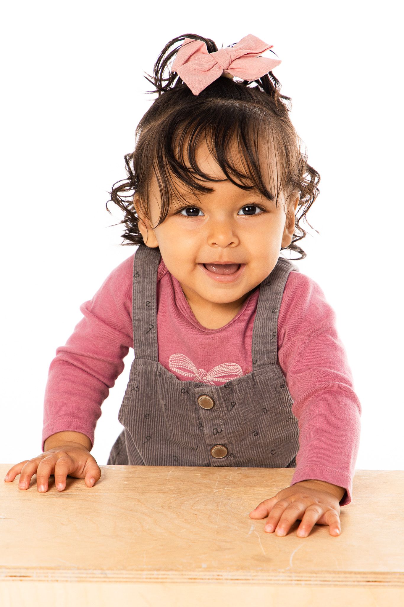 Toddler in pink shirt and gray overalls smiles while leaning on a wooden surface.