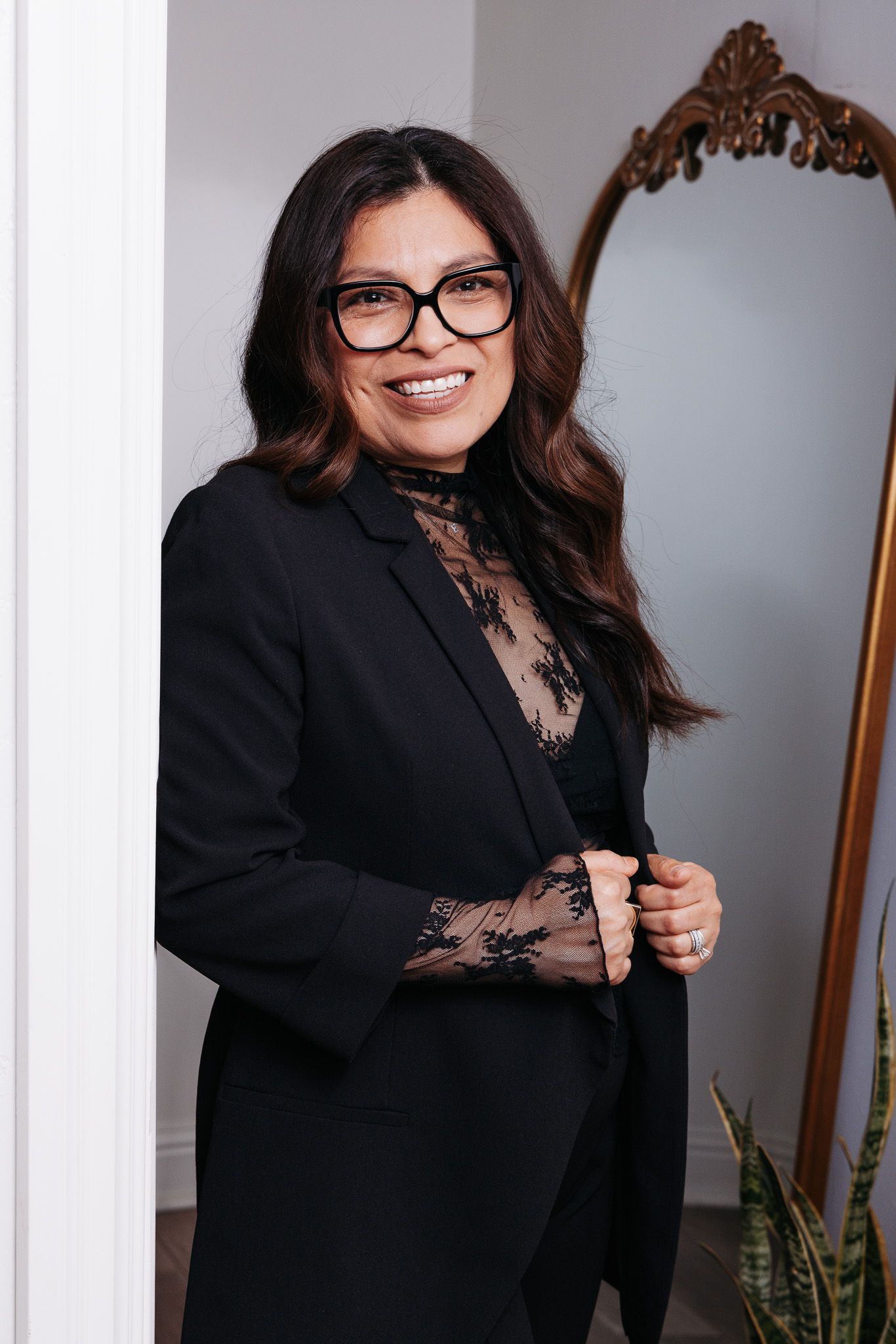 Professional wearing a black blazer and lace top with glasses poses elegantly against a wall with an ornate mirror.