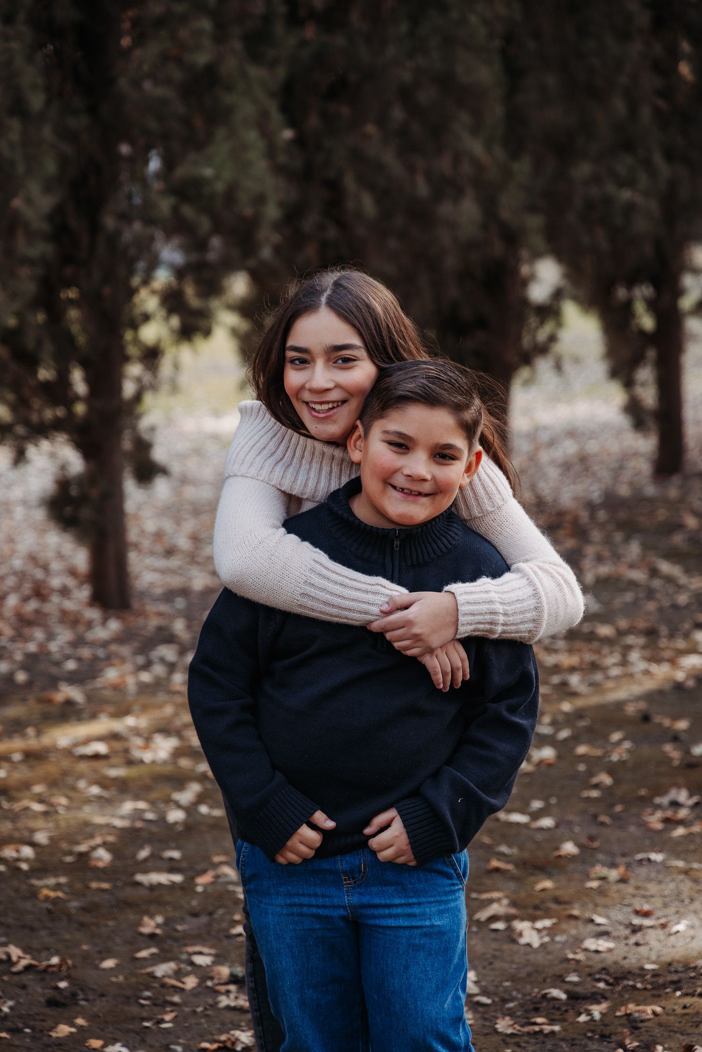 Two people in casual sweaters and jeans share playful hugs and laughs in a fall outdoor setting.