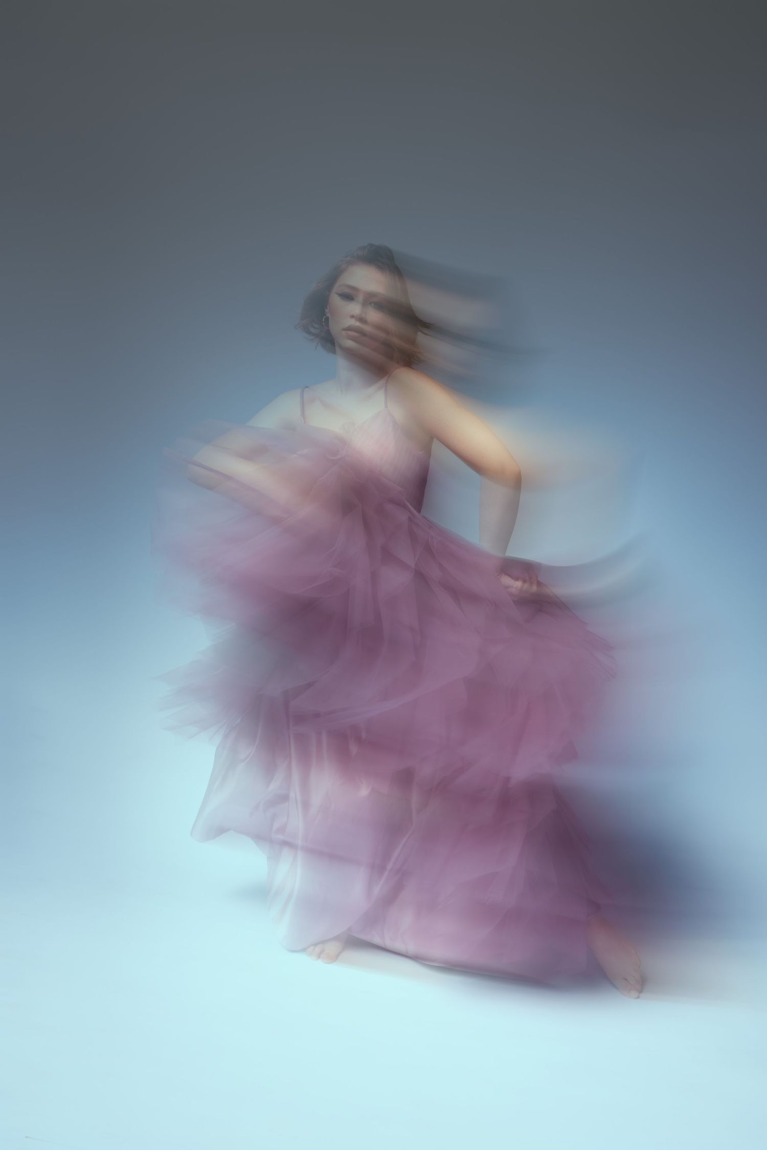 A dancer in a flowing mauve tulle dress creates an ethereal motion blur effect against a pale blue background.