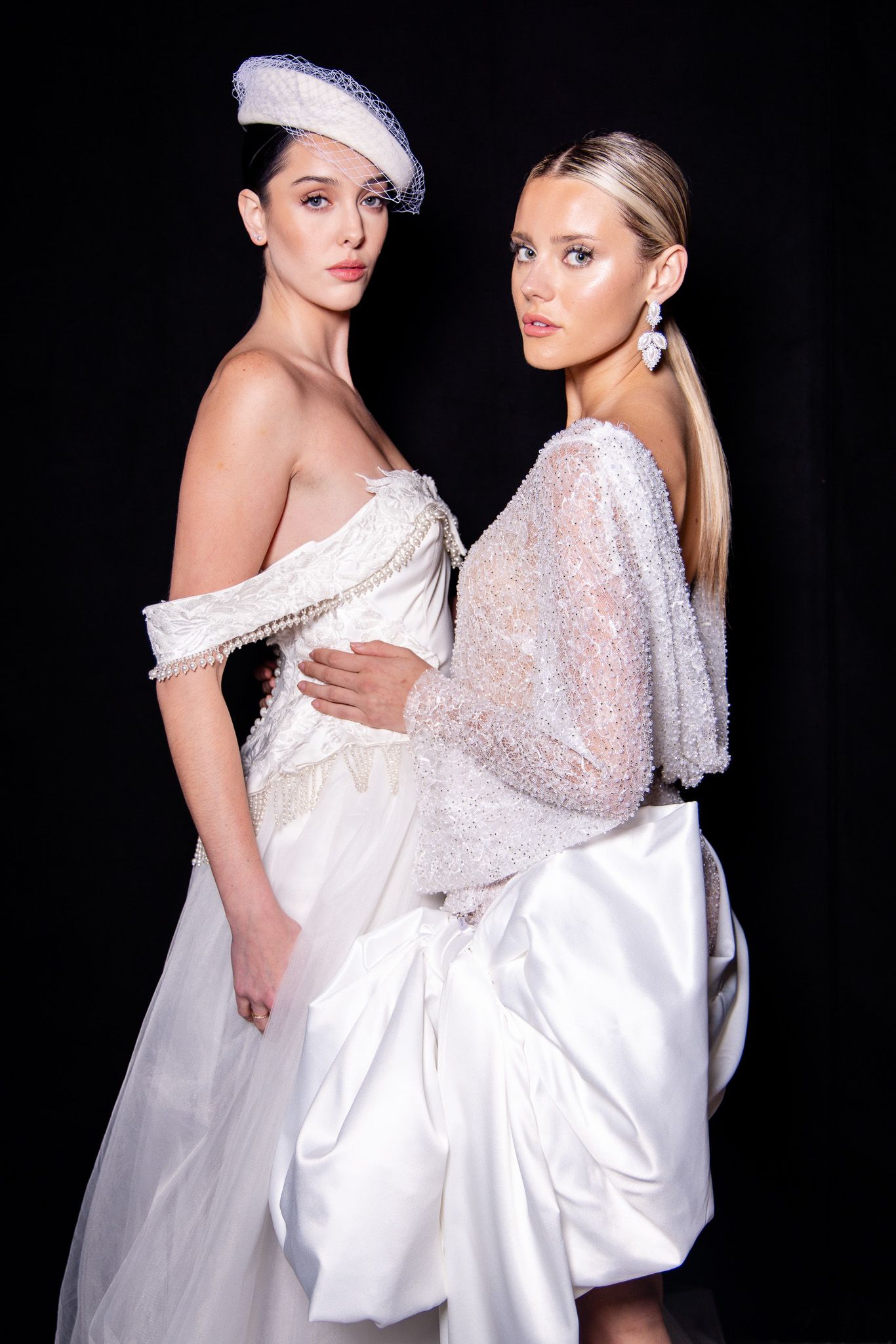 Models showcase elegant white bridal gowns with delicate lace details against a dark backdrop during a fashion show.
