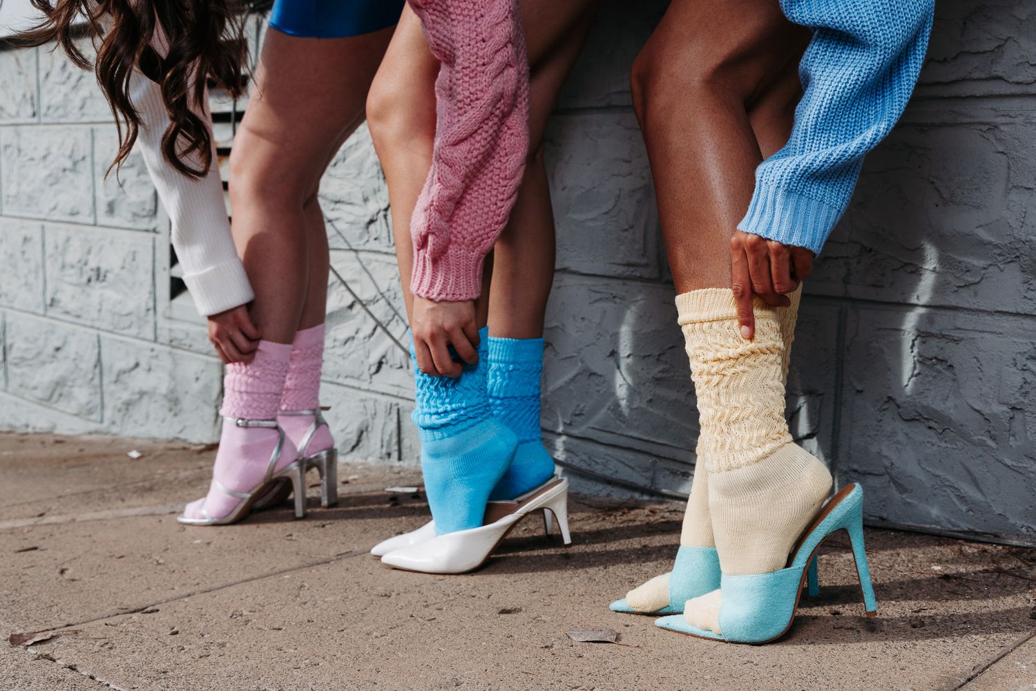 Three pairs of legs wearing colorful socks and high heels in blue, pink and cream colors against a gray wall.