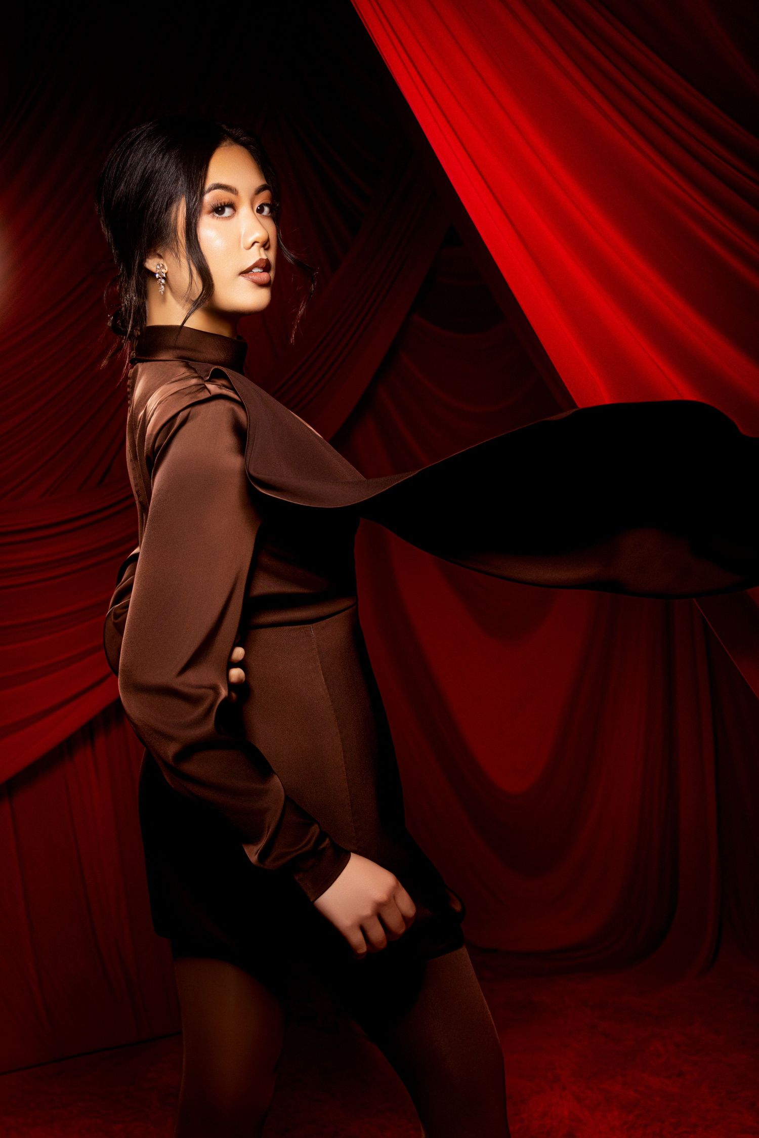 A dramatic fashion photoshoot against red velvet curtains featuring dark brown clothing and moody lighting.