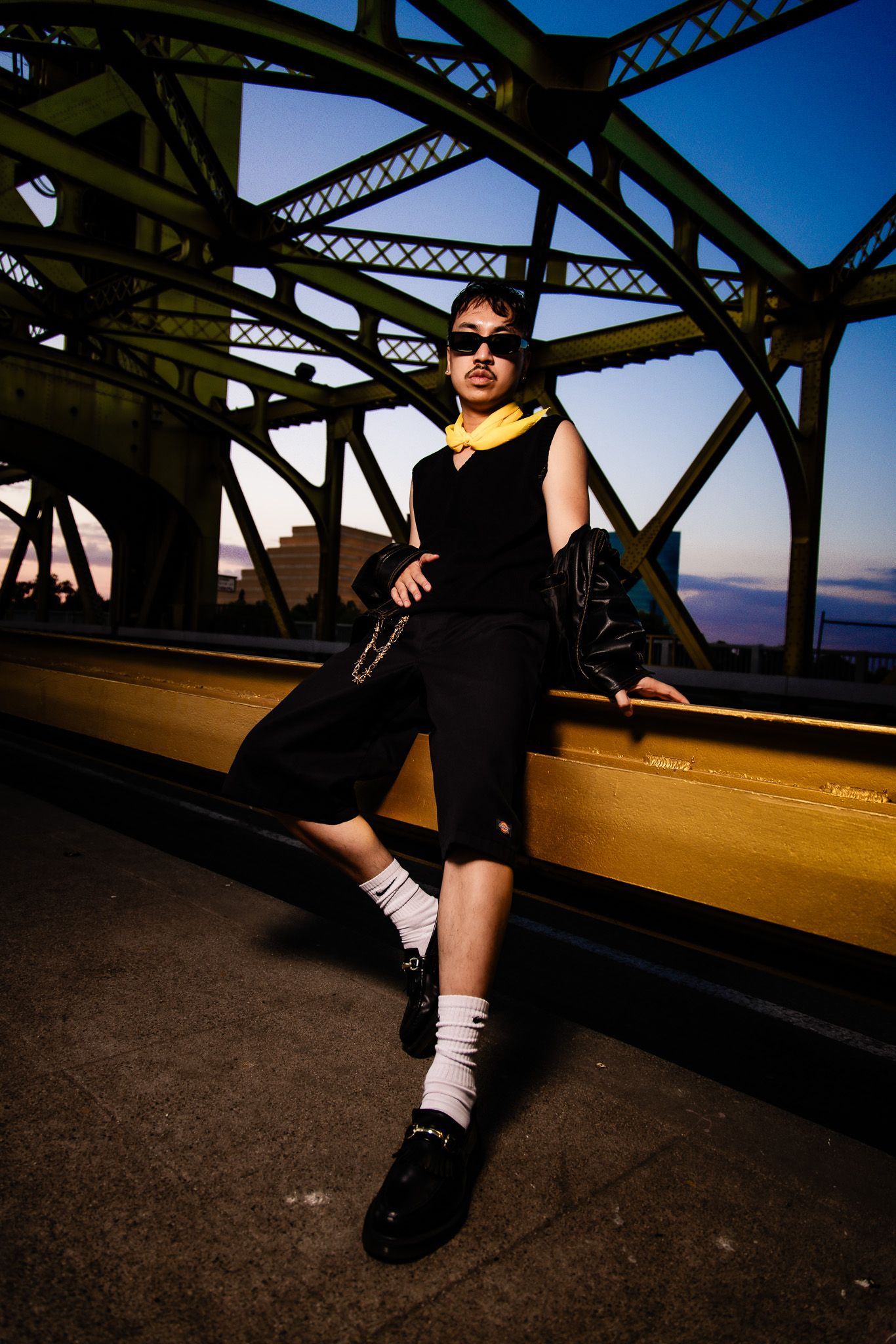 Person wearing black clothing and white socks reclines against urban bridge steel structure at dusk.