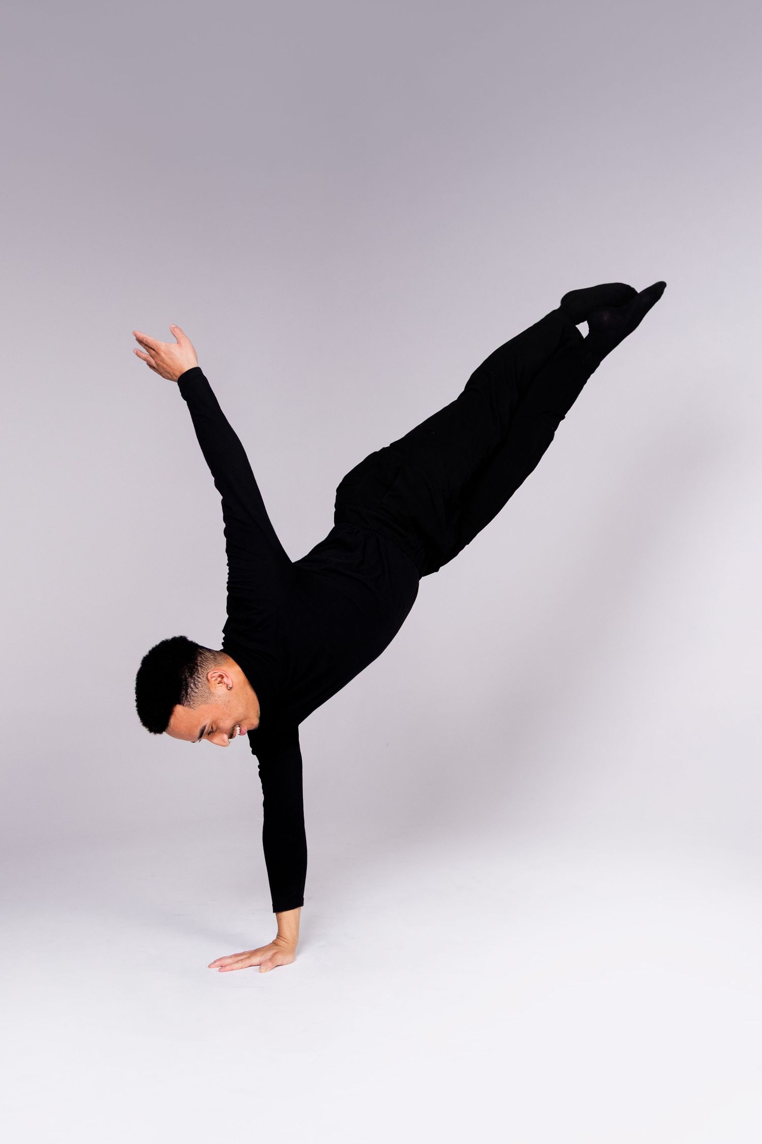 Dancer in black performs a one-handed handstand with extended leg against gray backdrop.