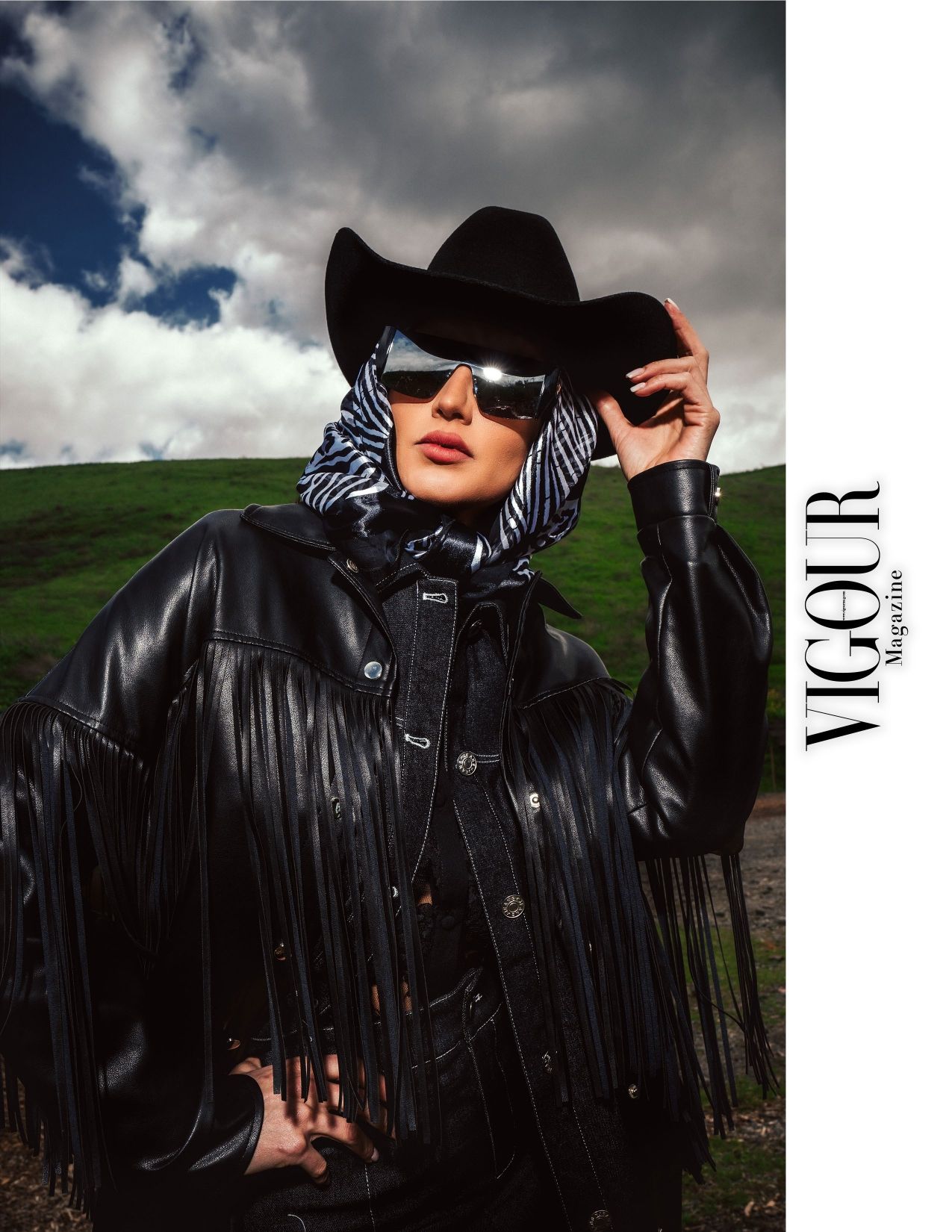 Model in black leather fringe jacket and cowboy hat poses against dramatic cloudy landscape.