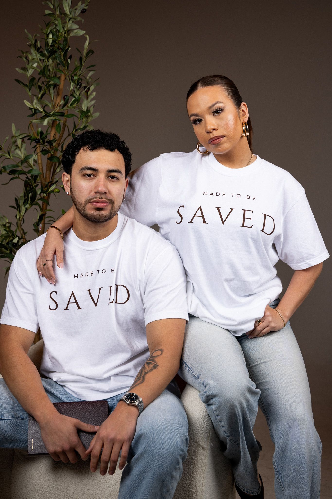 A series of lifestyle photos showing people wearing white 'SAVED' t-shirts and blue jeans in studio portraits against a brown backdrop.