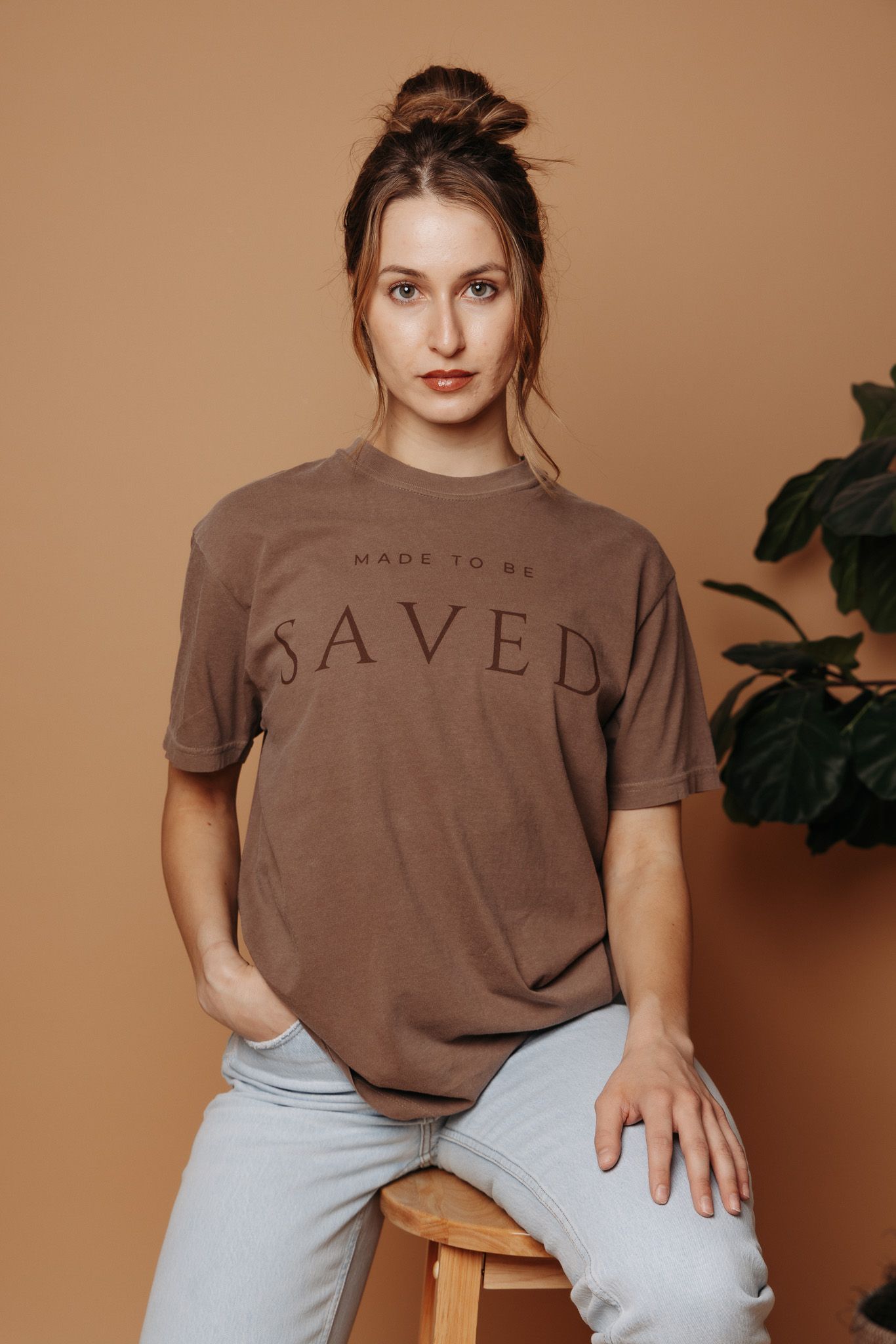 Casual brown t-shirt with 'SAVED' text paired with light denim jeans against a neutral brown background with plant decor.