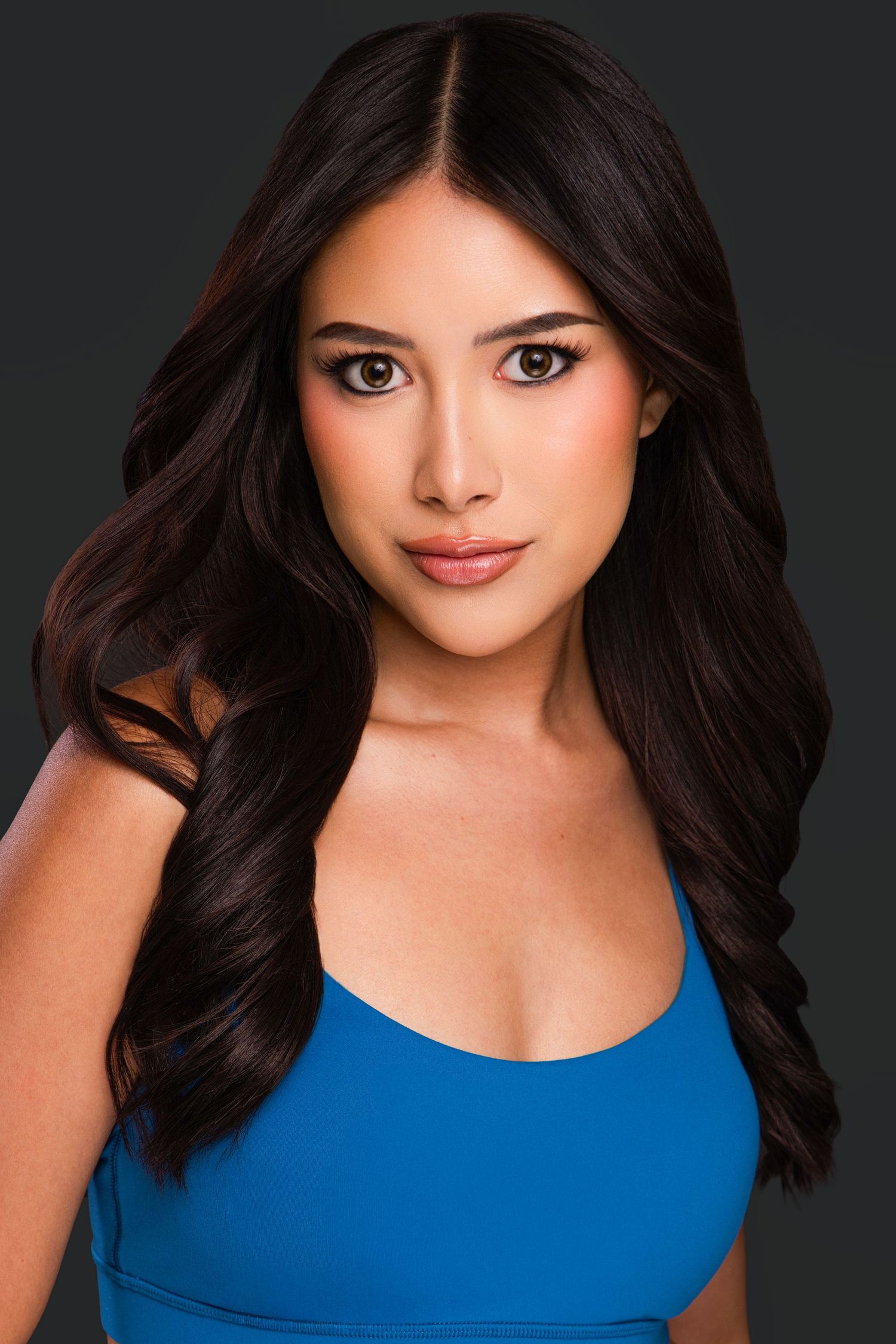 Young woman with long dark wavy hair wearing a blue top poses for a professional headshot portrait.