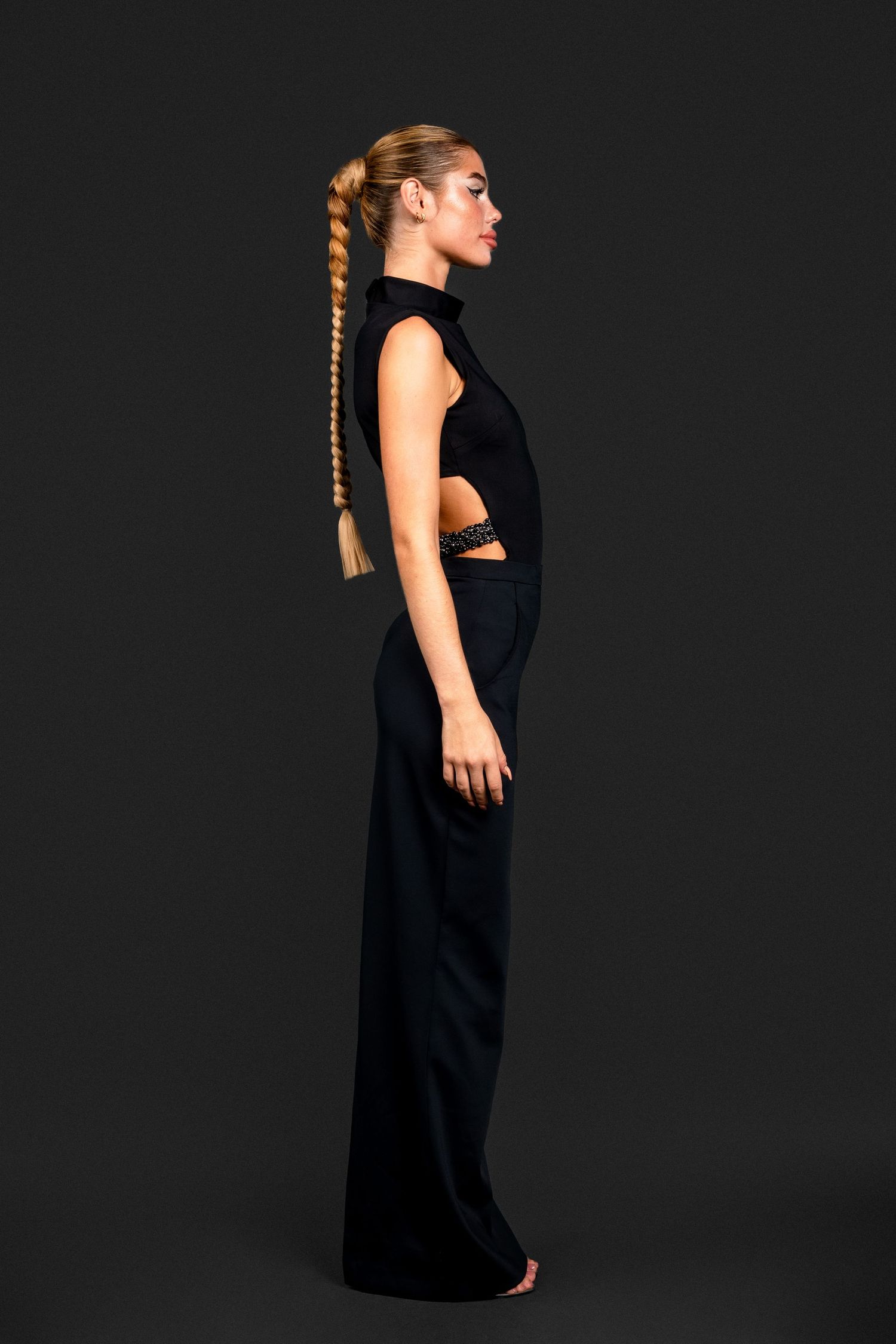 Model in profile wearing a black sleeveless cutout floor-length gown with a long braided ponytail hairstyle.