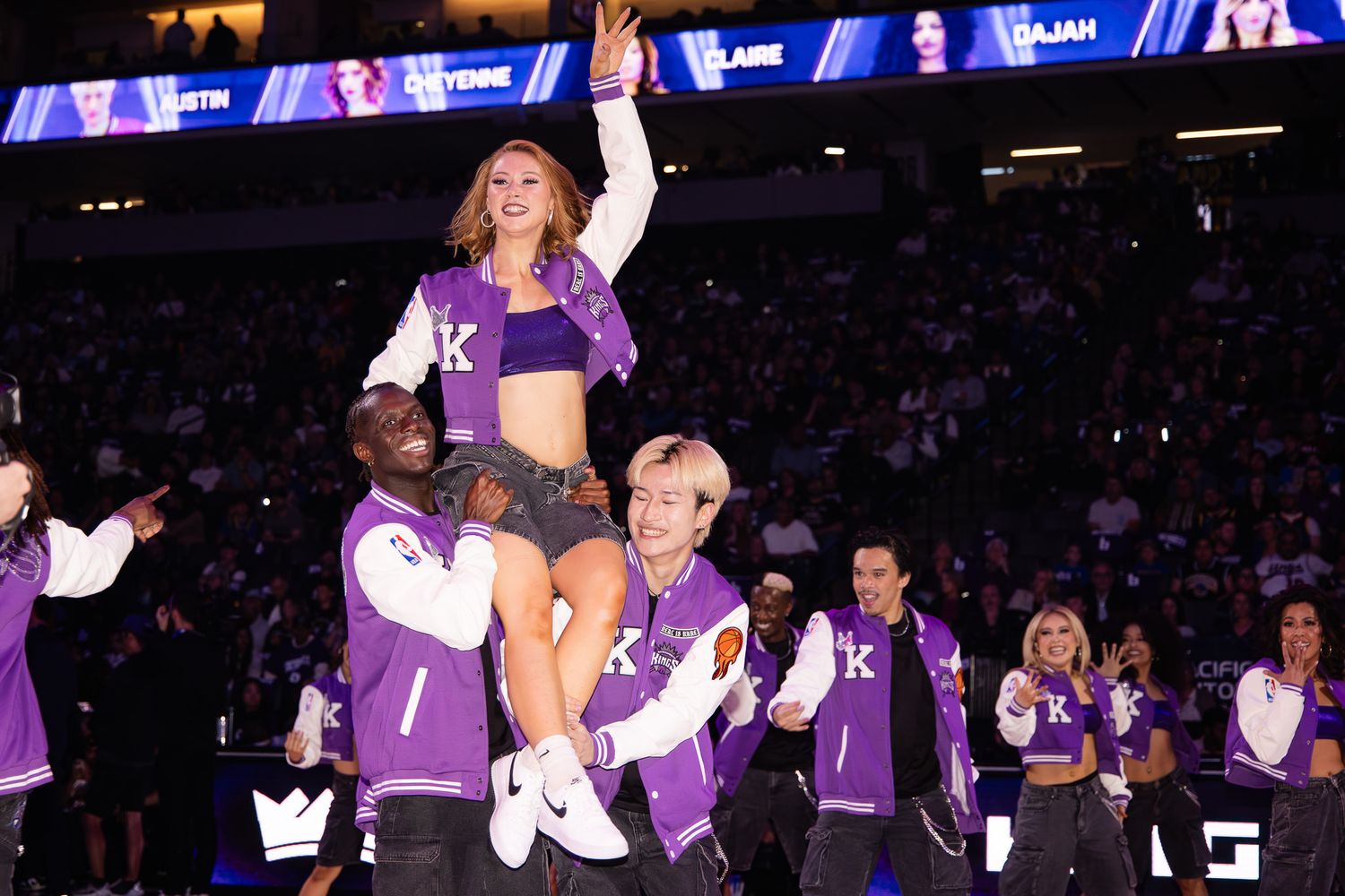 Cheerleaders in purple jackets perform a lift routine at an indoor basketball arena during a game.