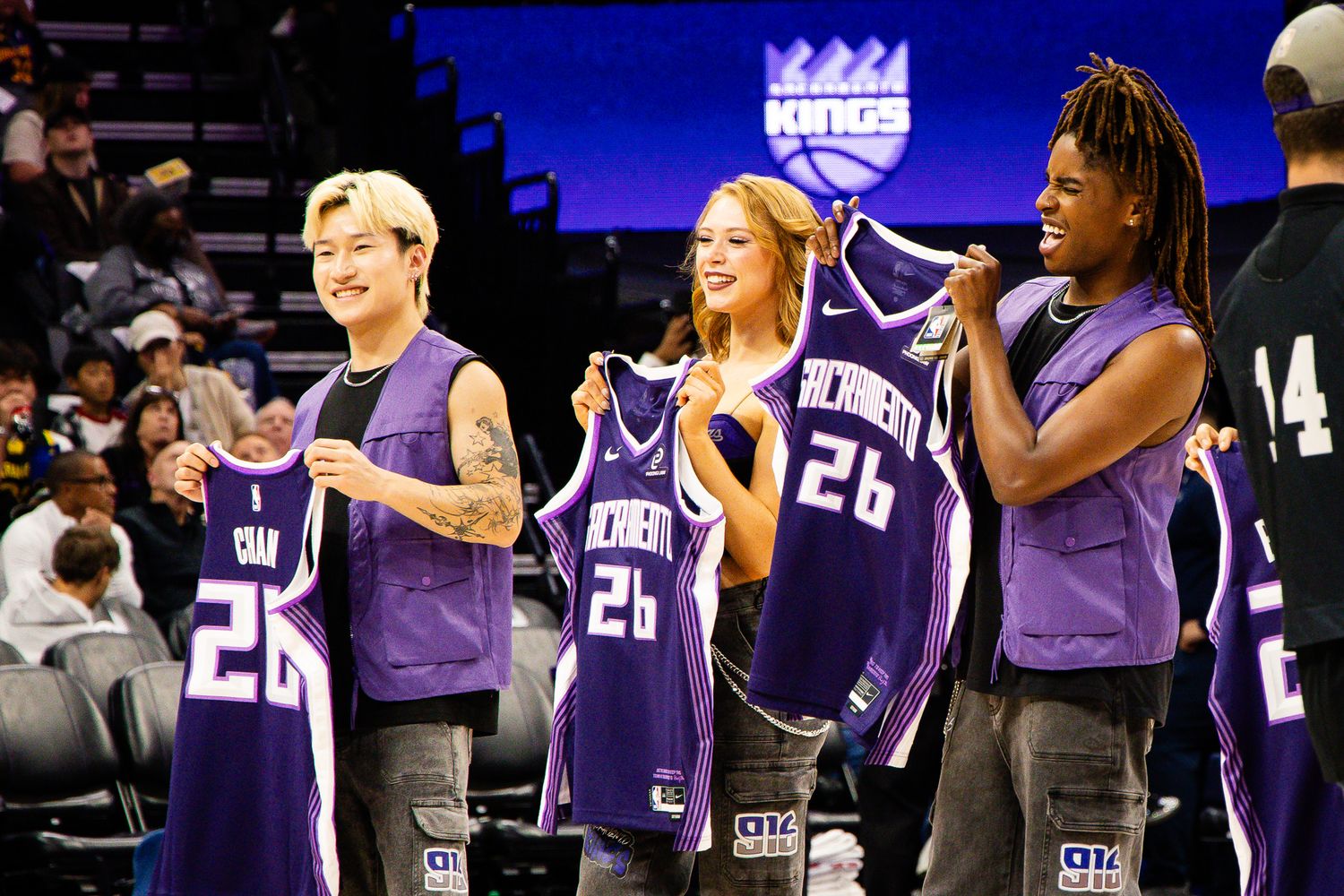 Three fans smile holding Sacramento Kings purple jerseys numbered 26 at an NBA arena event.
