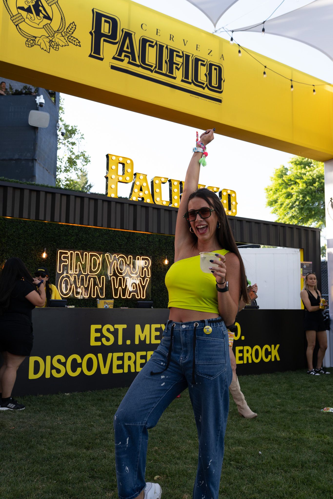 Festival attendee in yellow crop top and jeans poses at Pacifico beer branded event with neon sign reading 'Find Your Own Way'.