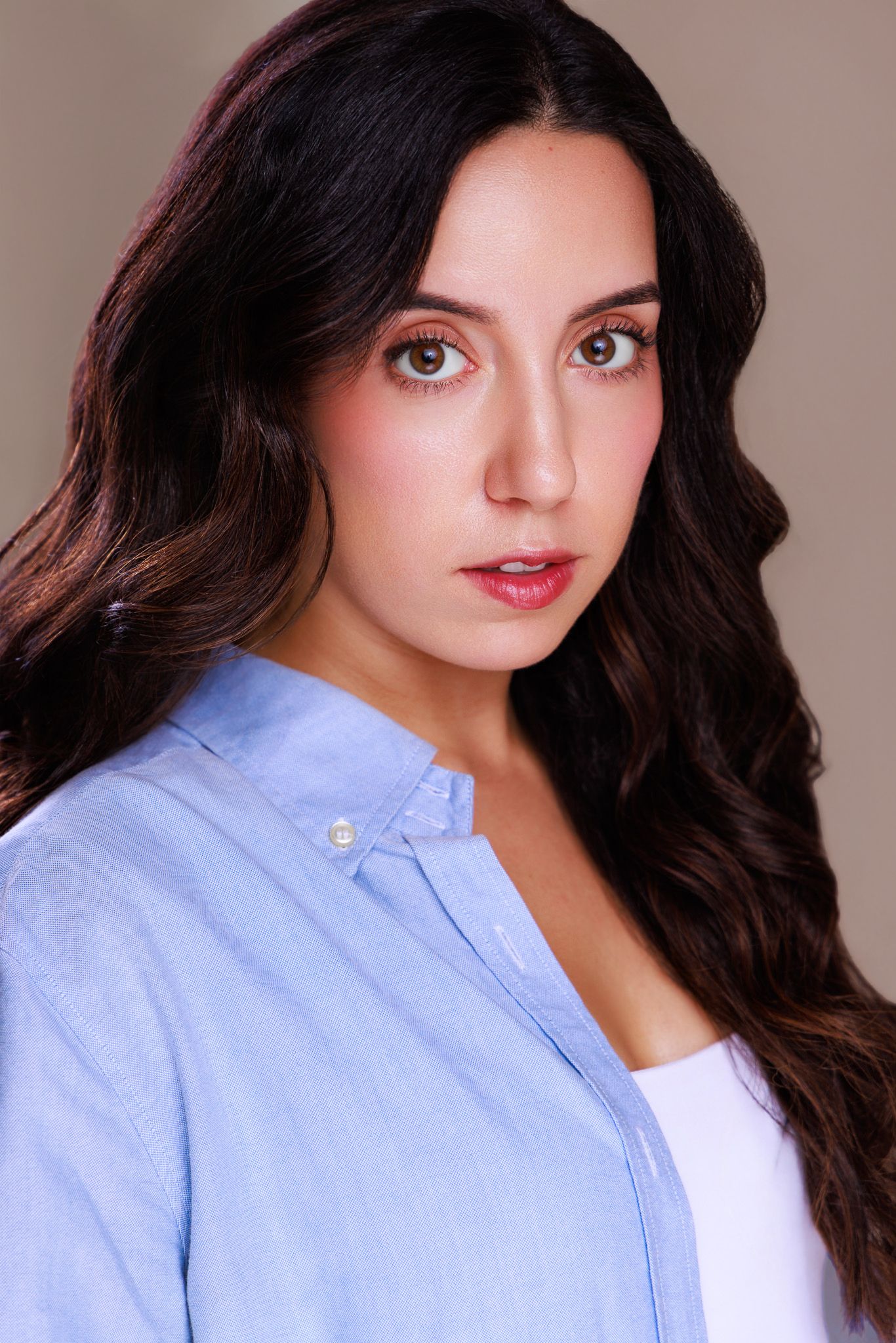 Young woman with long wavy dark hair wearing a blue button-up shirt poses for a professional headshot photo.
