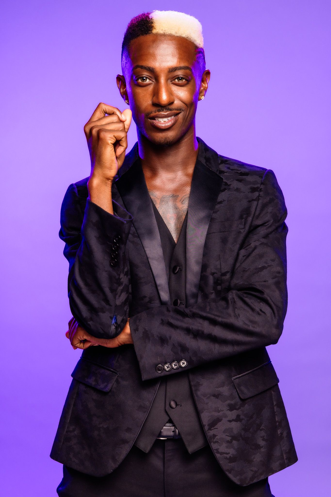 Stylish young Black man with blonde hair wearing a black velvet suit, posing confidently against a purple background.