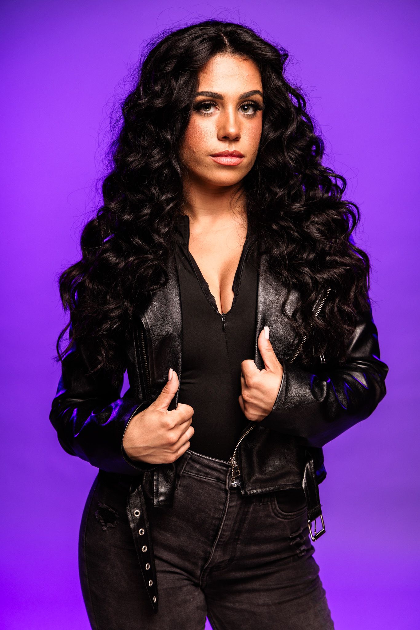Woman with long curly black hair wearing a black leather jacket and jeans poses against a purple background.
