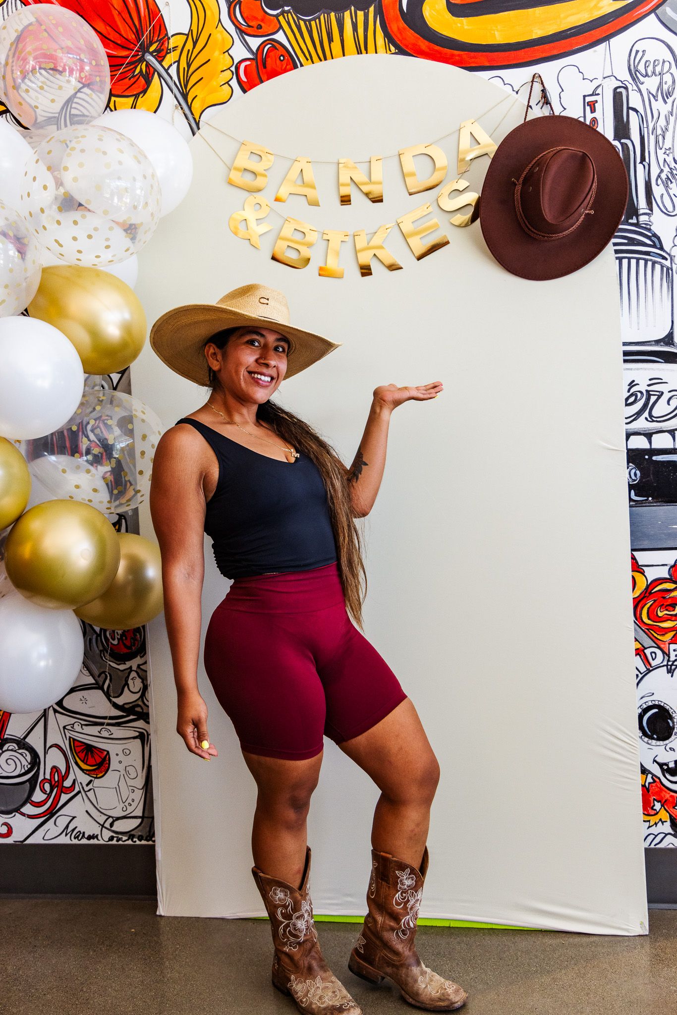 Woman in cowboy hat and boots poses at Banda & Bikes party with gold balloons.
