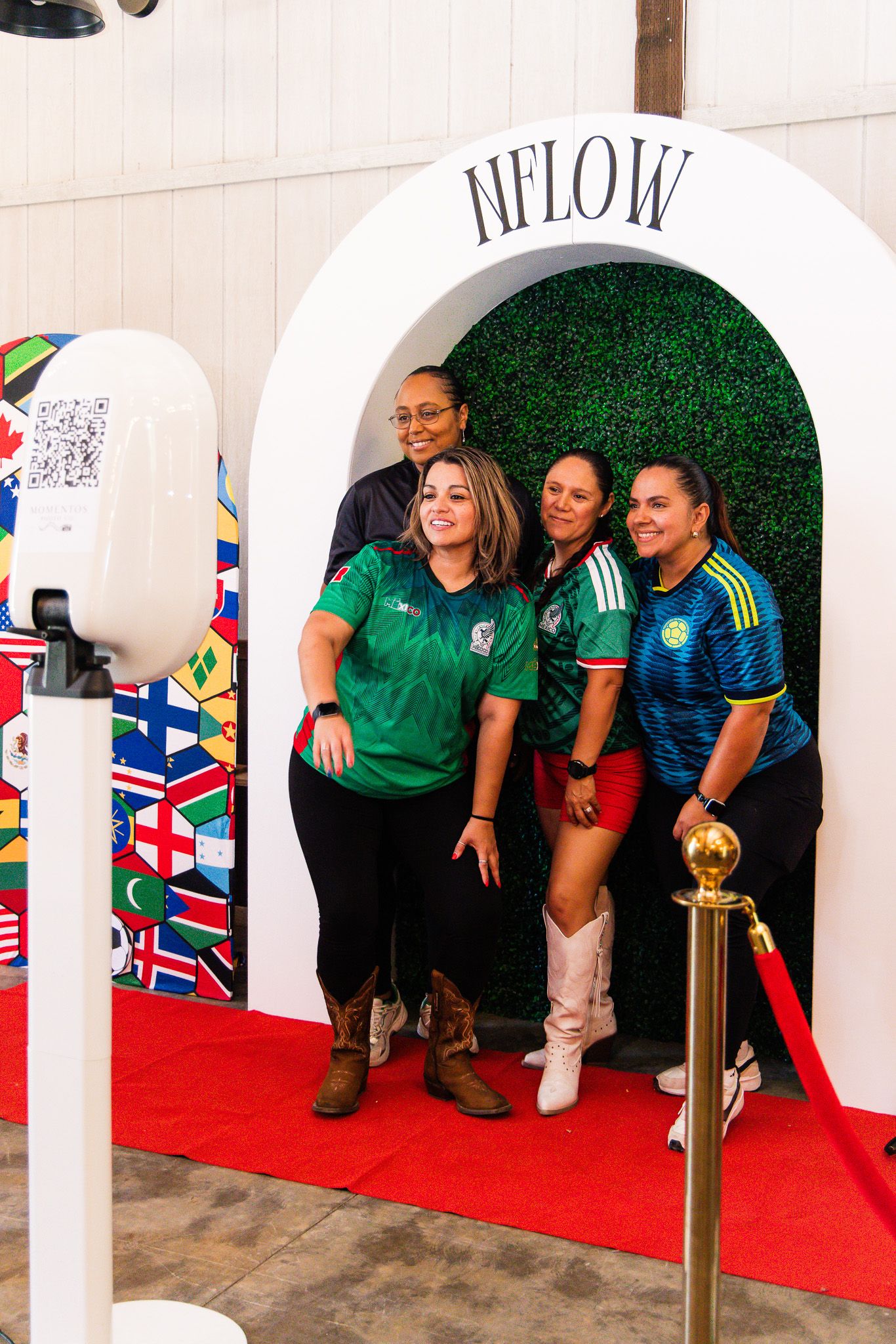 Four women posing together under an NFLOW arch with green hedge backdrop on a red carpet at an event.