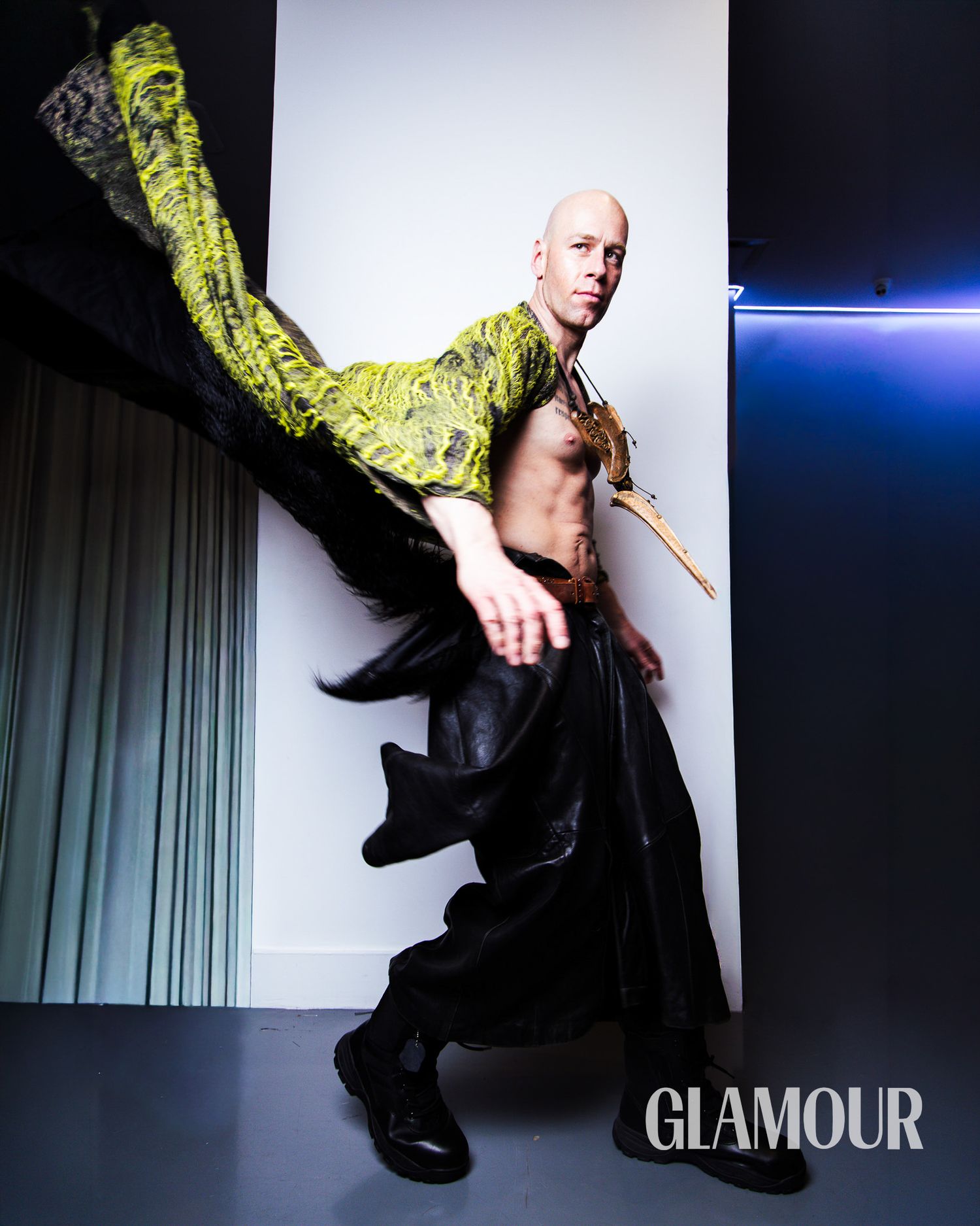 Bald male model poses dynamically in green patterned jacket and black skirt for Glamour magazine editorial.