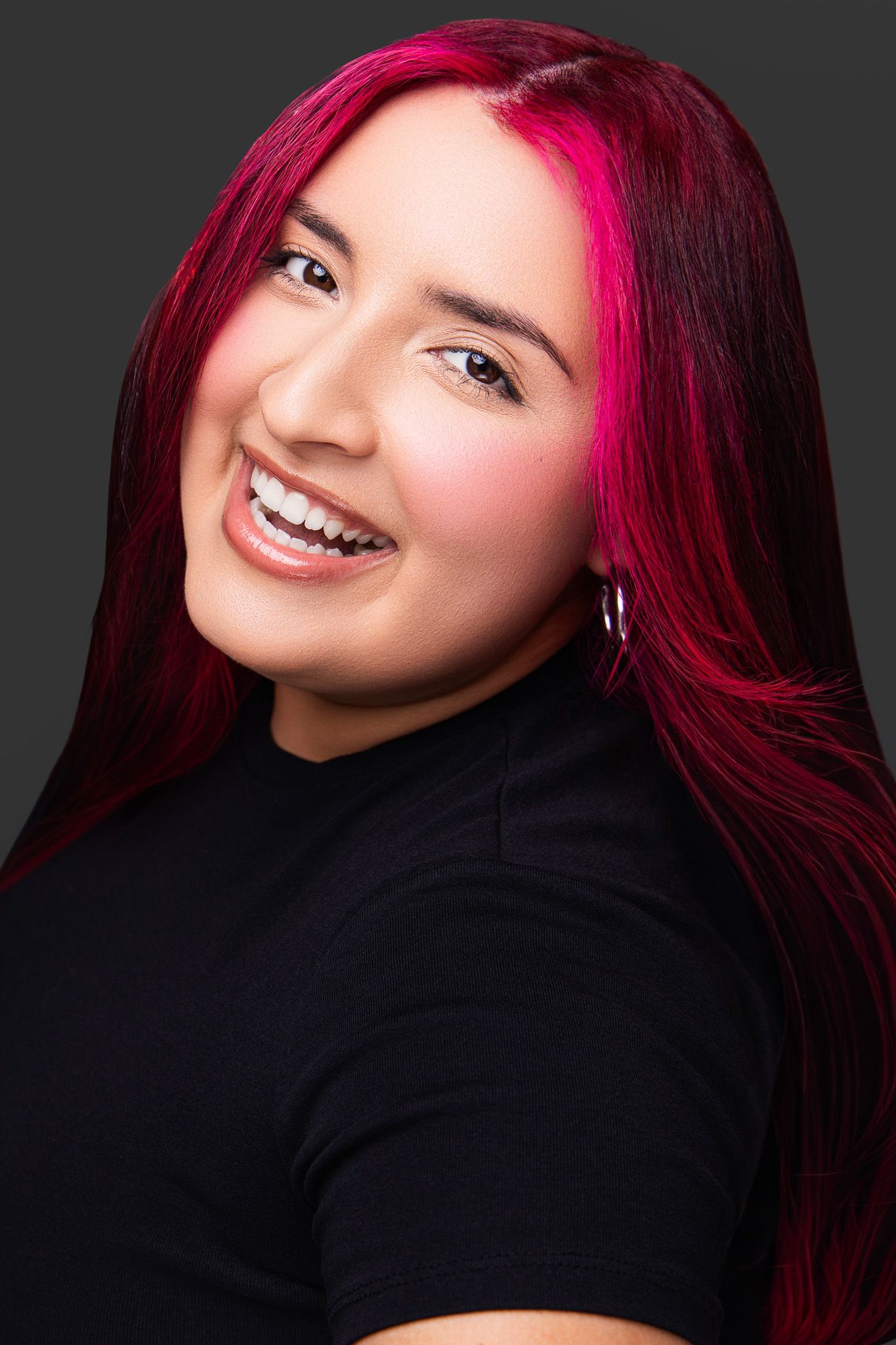 Smiling young woman with vibrant pink and red hair wearing a black top against a dark gray background.