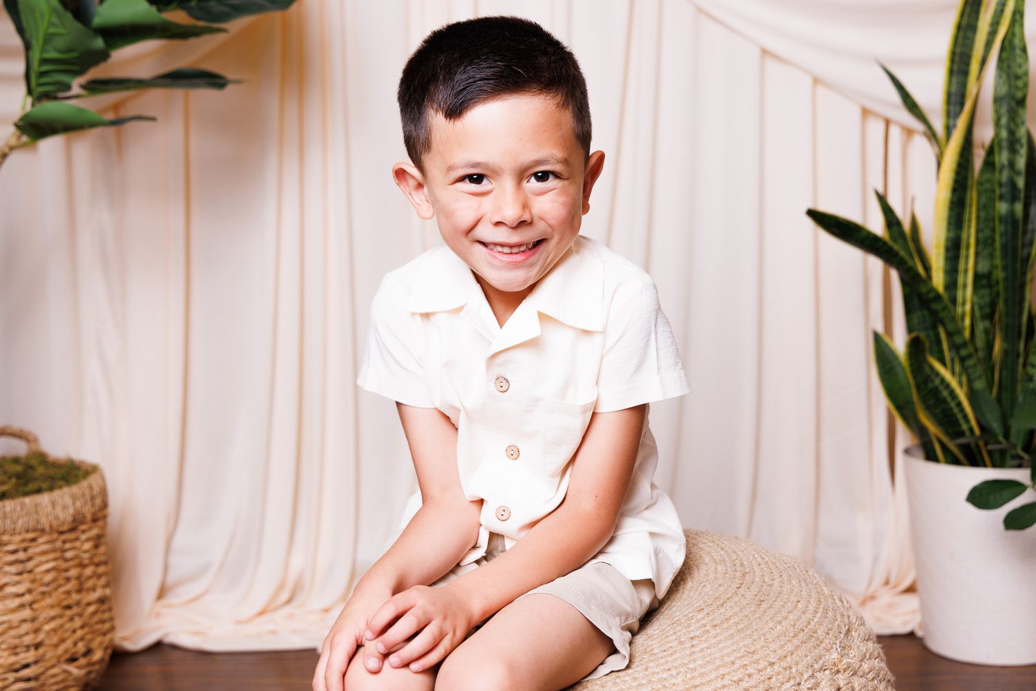 Smiling young boy in white romper sitting on wicker ottoman surrounded by green plants and neutral backdrop.