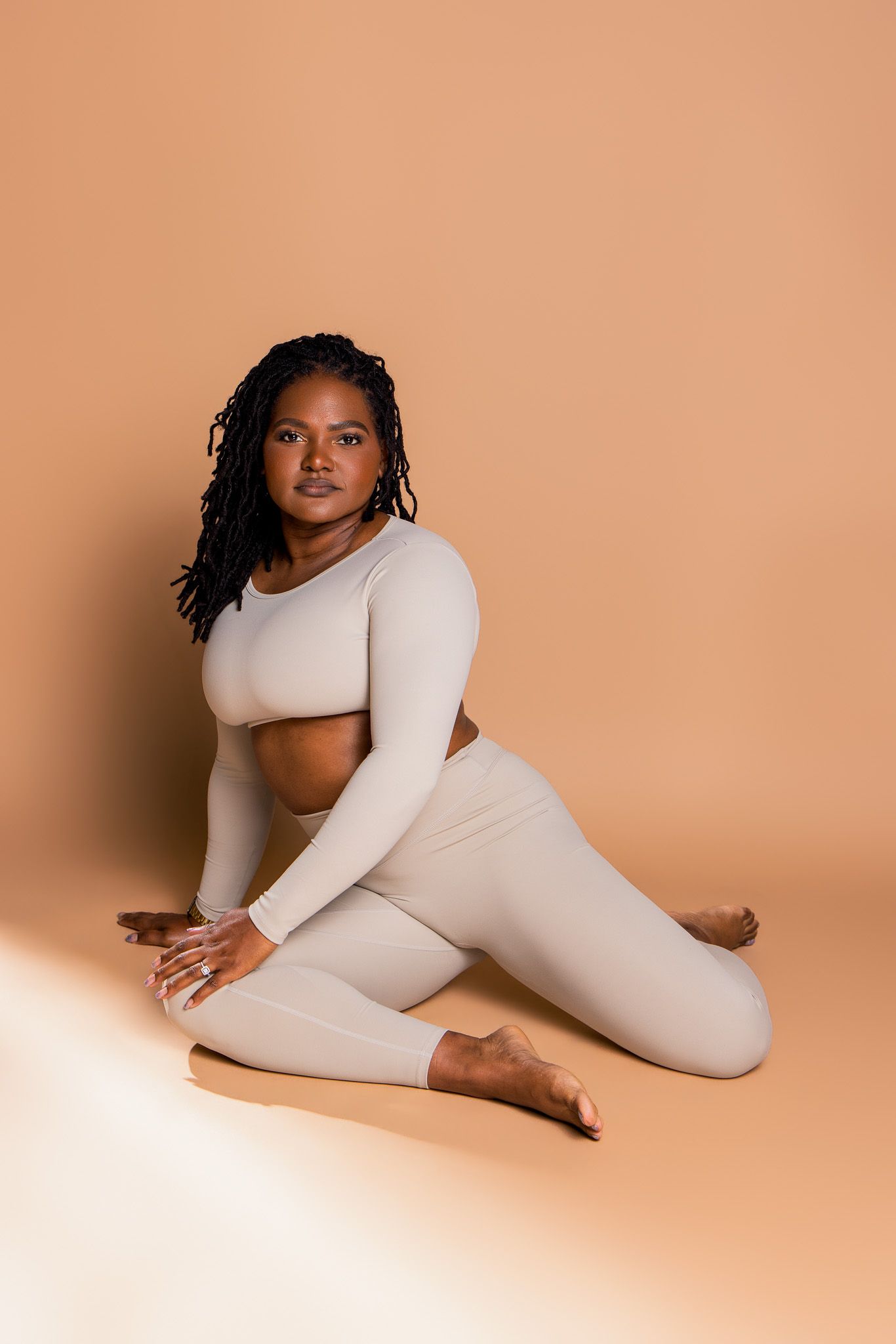 Woman in beige long-sleeve crop top and leggings sitting gracefully on a beige studio floor.