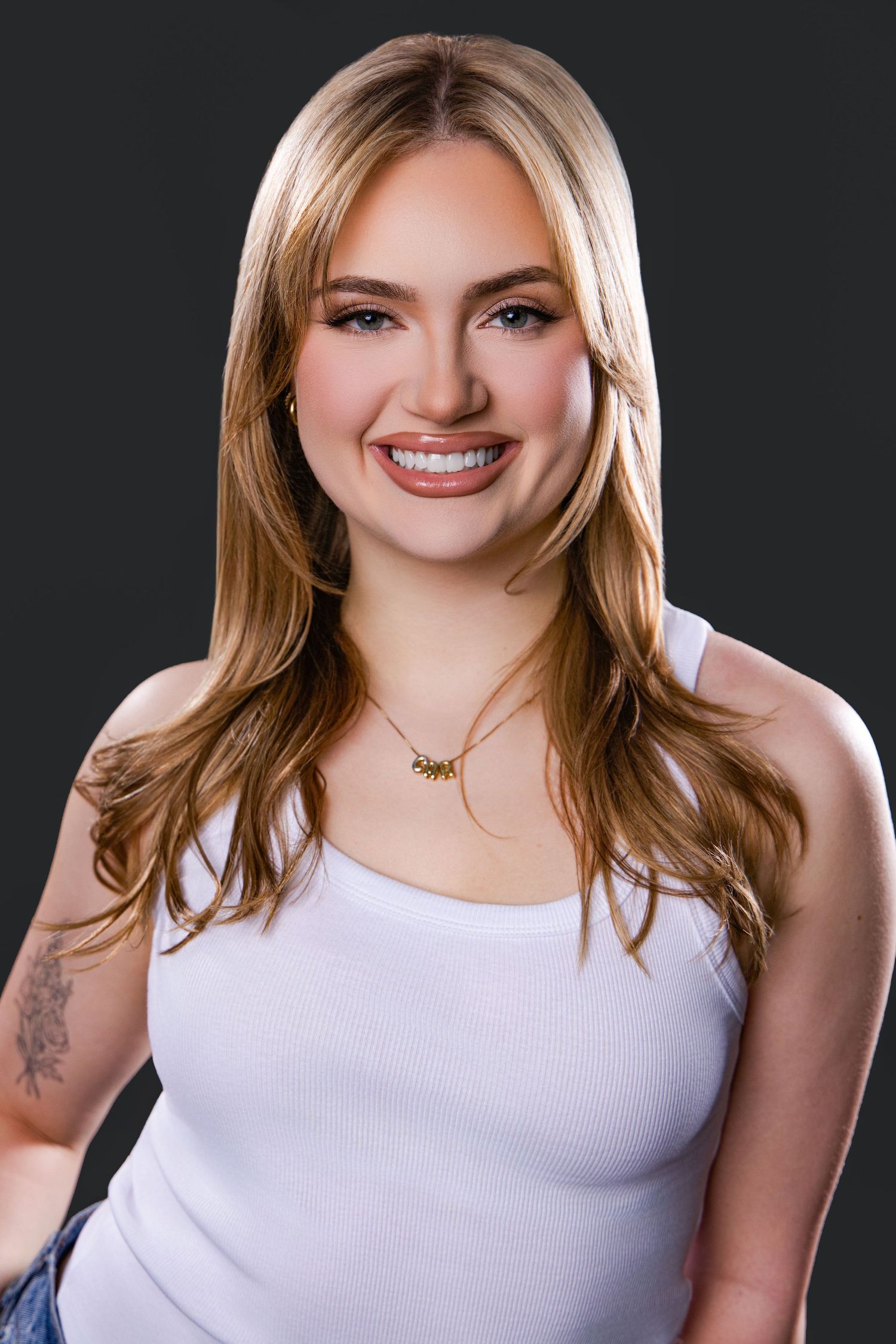 Smiling young blonde woman with tattoo wearing white tank top and gold necklace against dark gray background.
