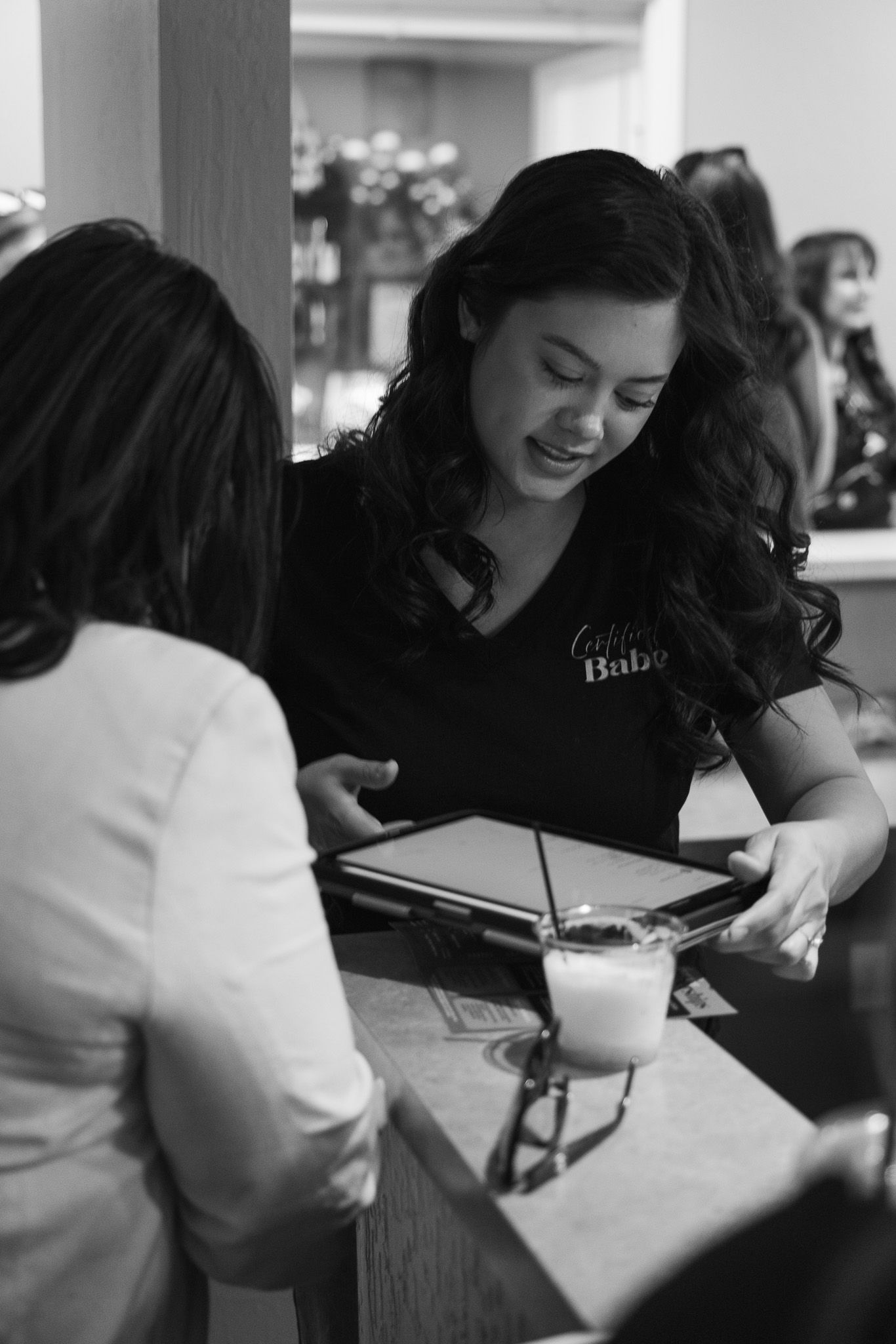 A smiling waitress in a black uniform shows a tablet menu to a customer at a restaurant counter in black and white.