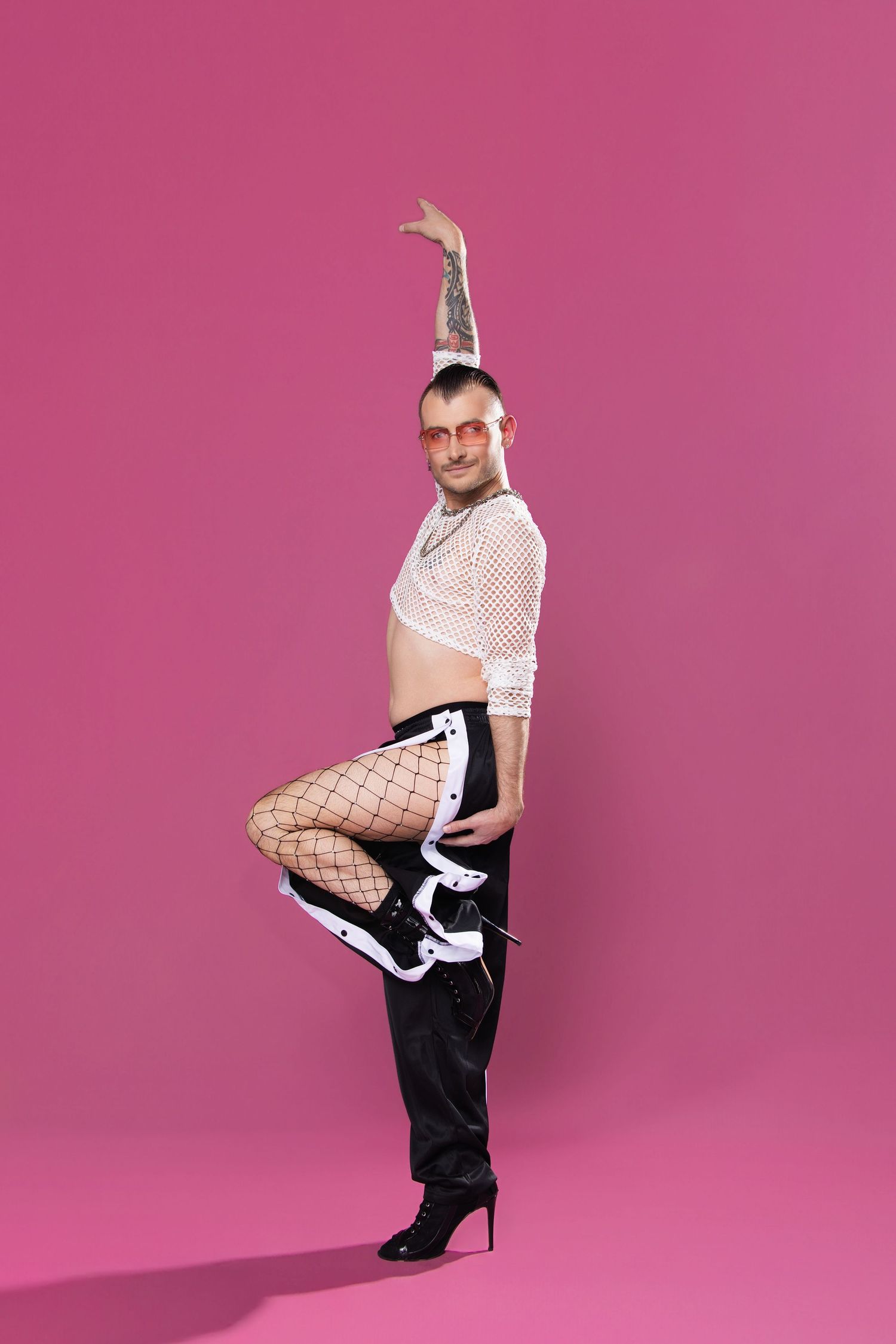 Man in mesh top, fishnet tights, black shorts and heels poses dramatically on pink background.