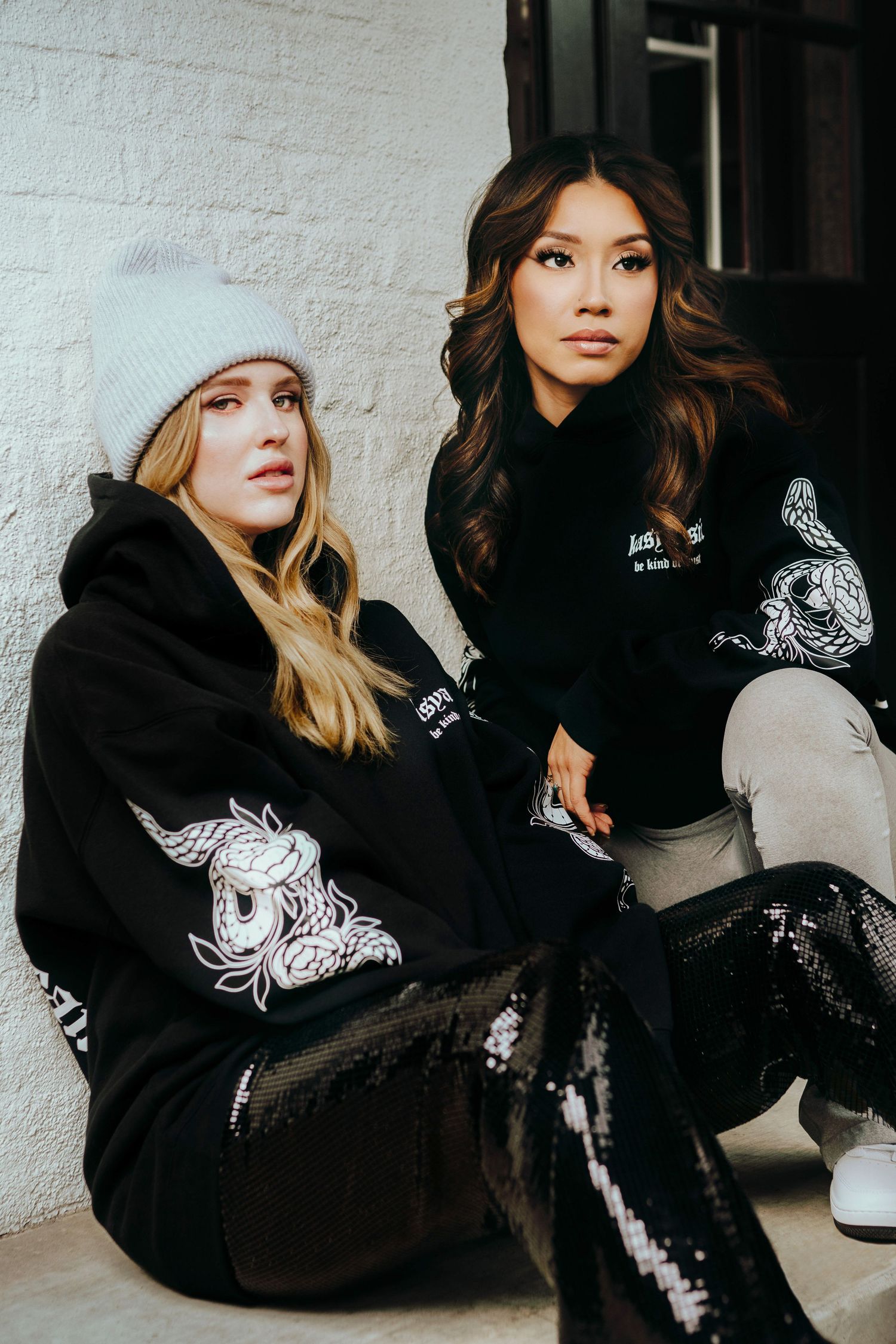 Two women in matching black graphic hoodies pose together, one wearing a gray beanie, against a white brick wall.