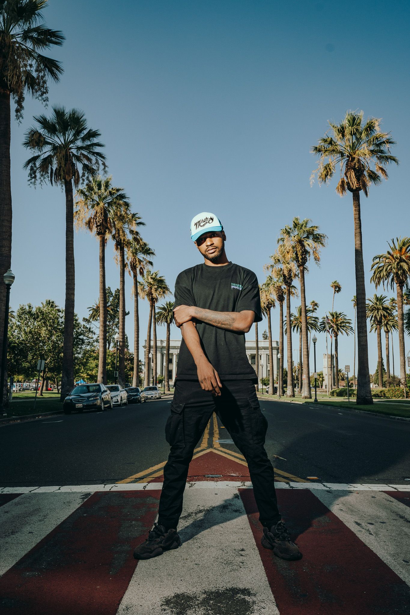 Young man in black outfit and white cap poses confidently on a crosswalk lined with tall palm trees under a clear blue sky.