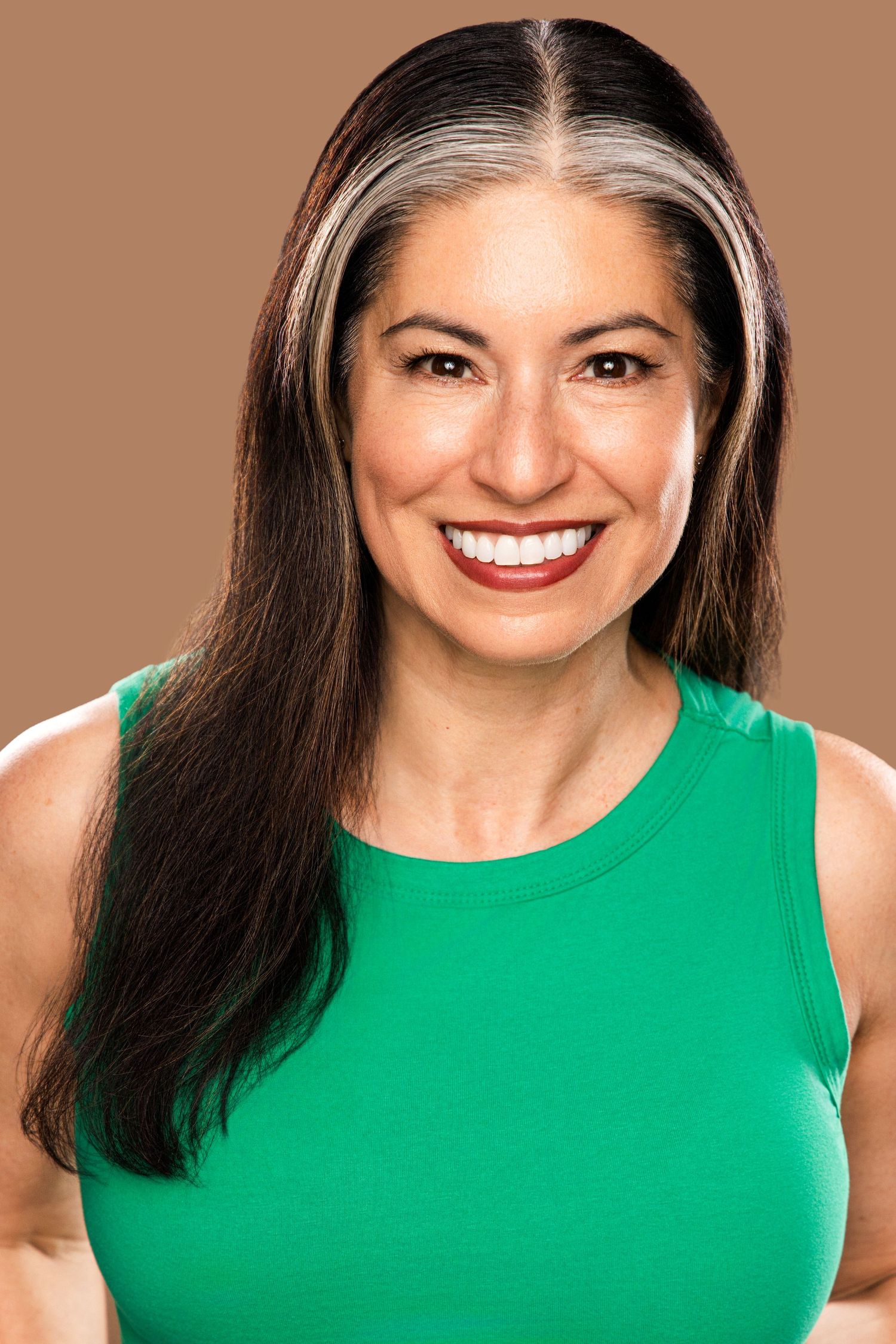 Smiling woman with dark hair and gray streaks wearing a green sleeveless top against a brown background.