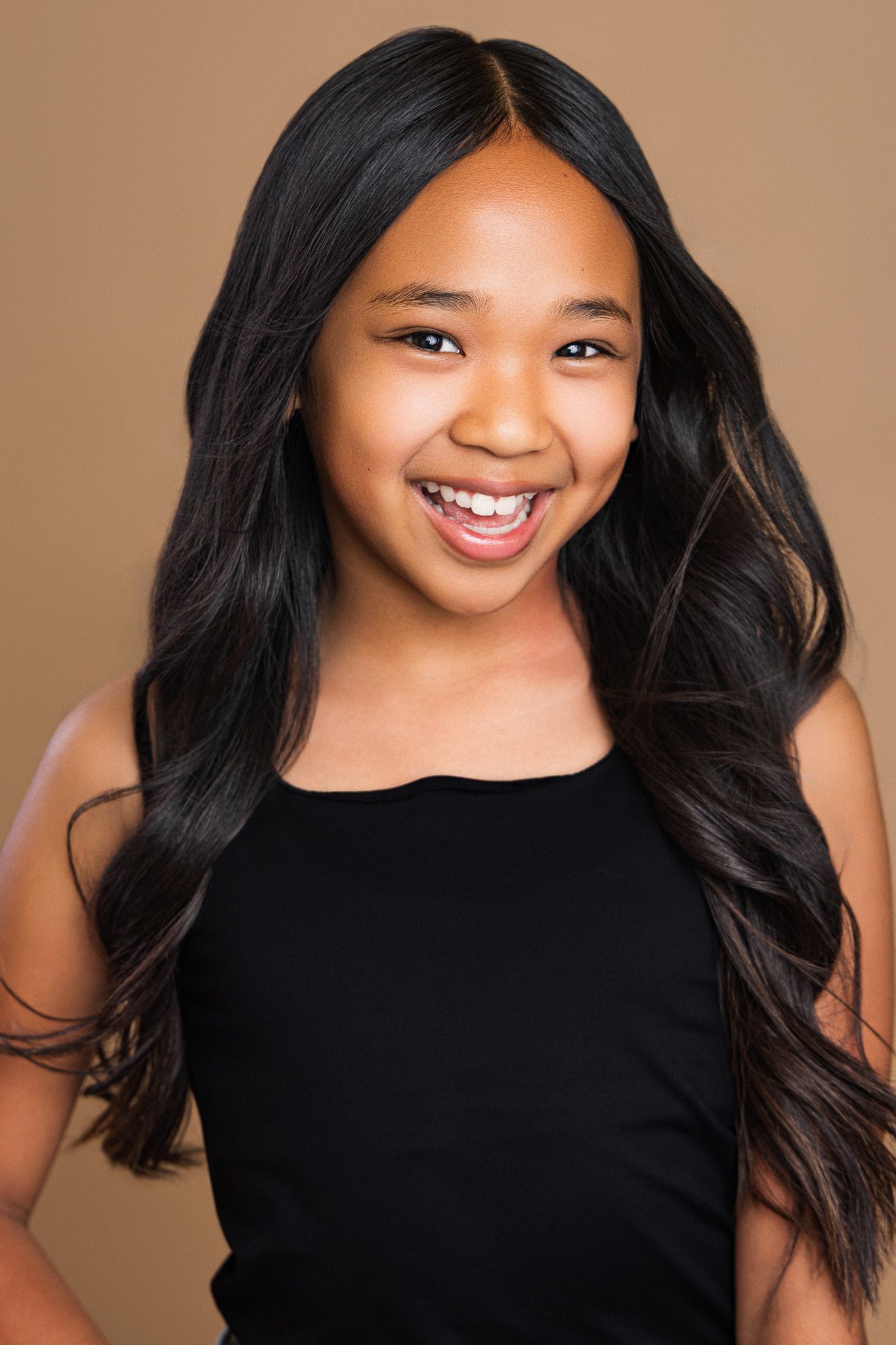 A young girl with long black wavy hair smiles brightly, wearing a black top against a brown background.