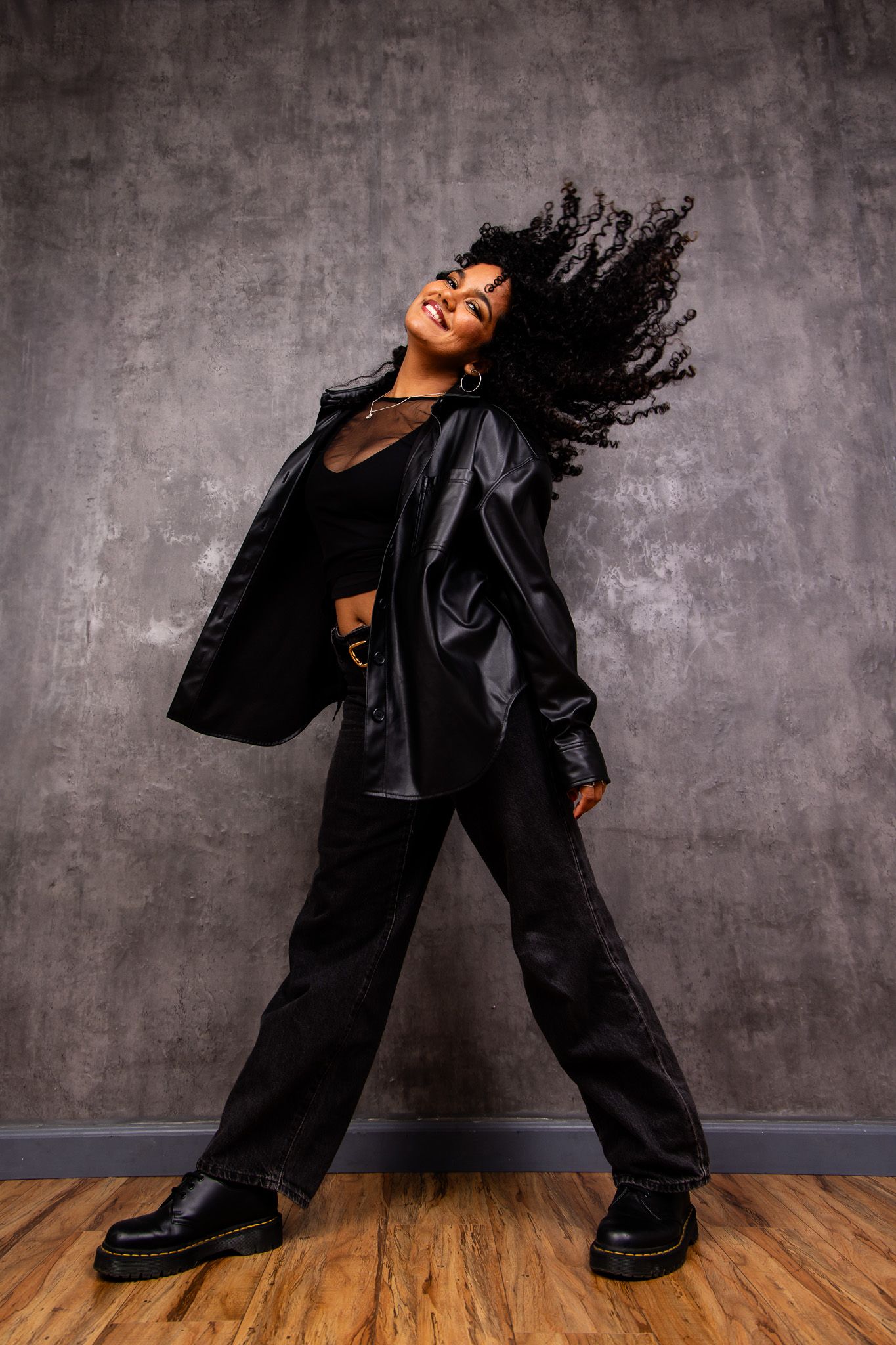 Energetic dancer in all black ensemble whips hair while striking a powerful pose on wooden floor.