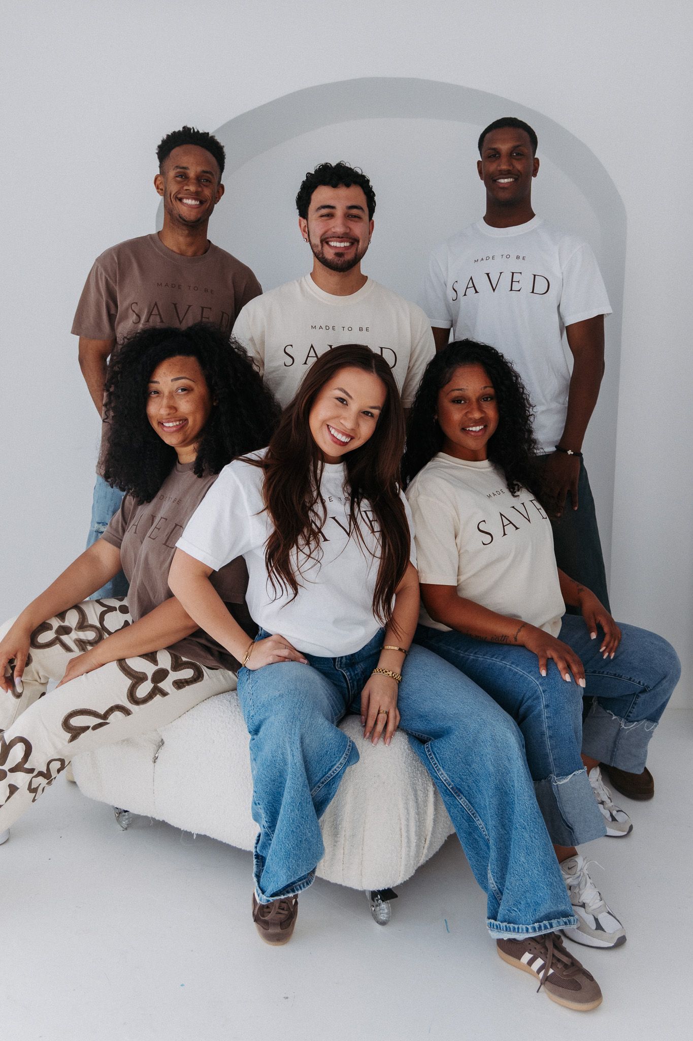 Friends laughing and posing together on a white couch wearing matching SAVED t-shirts and blue jeans.